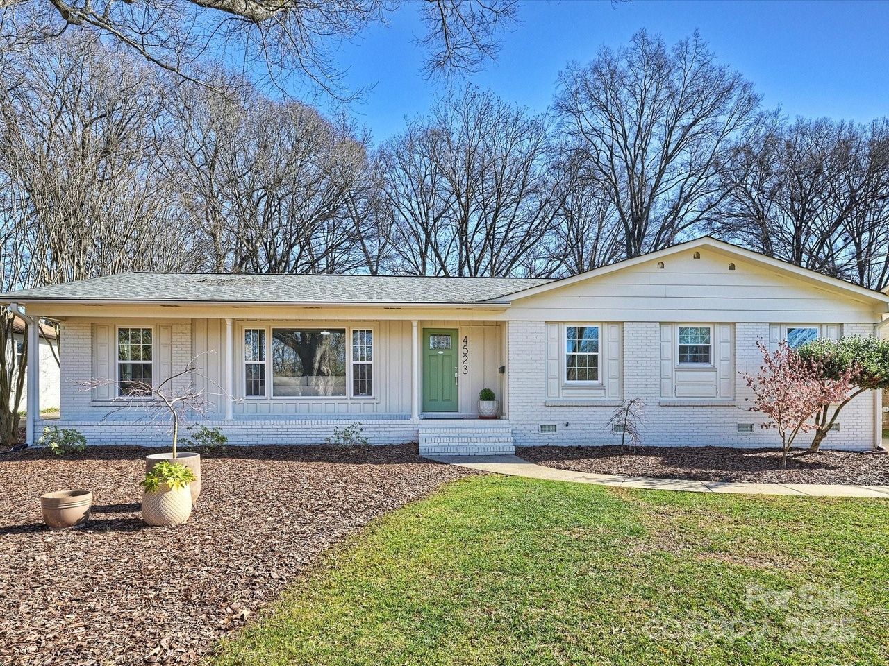 Single-story white brick house with a green front door, several windows, leafless trees in the background, and a front yard with mulch, potted plants, and a mix of grass and brown ground cover.
