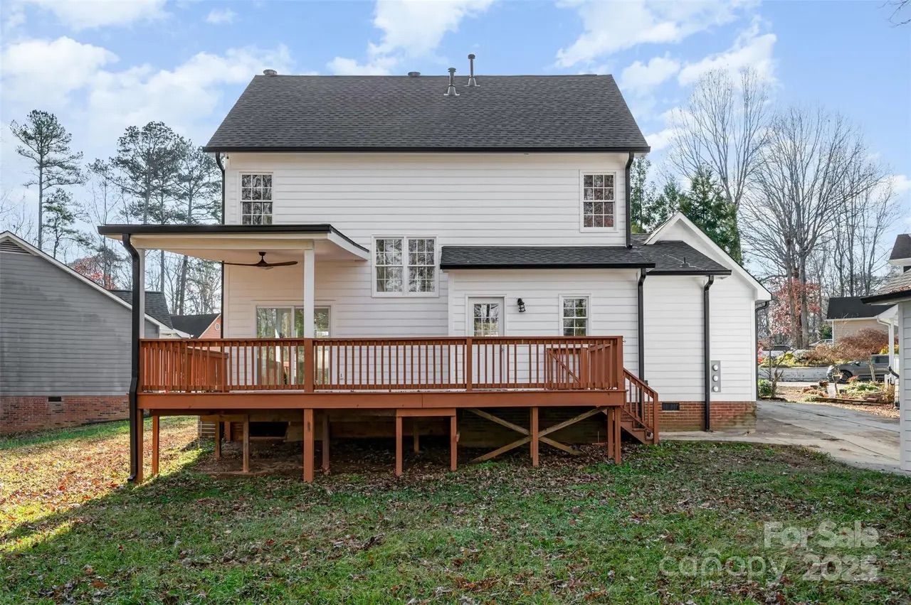 Rear view of a two-story white house with black roof, featuring a large wooden deck with railing and stairs leading to a grassy backyard under a partly cloudy sky.