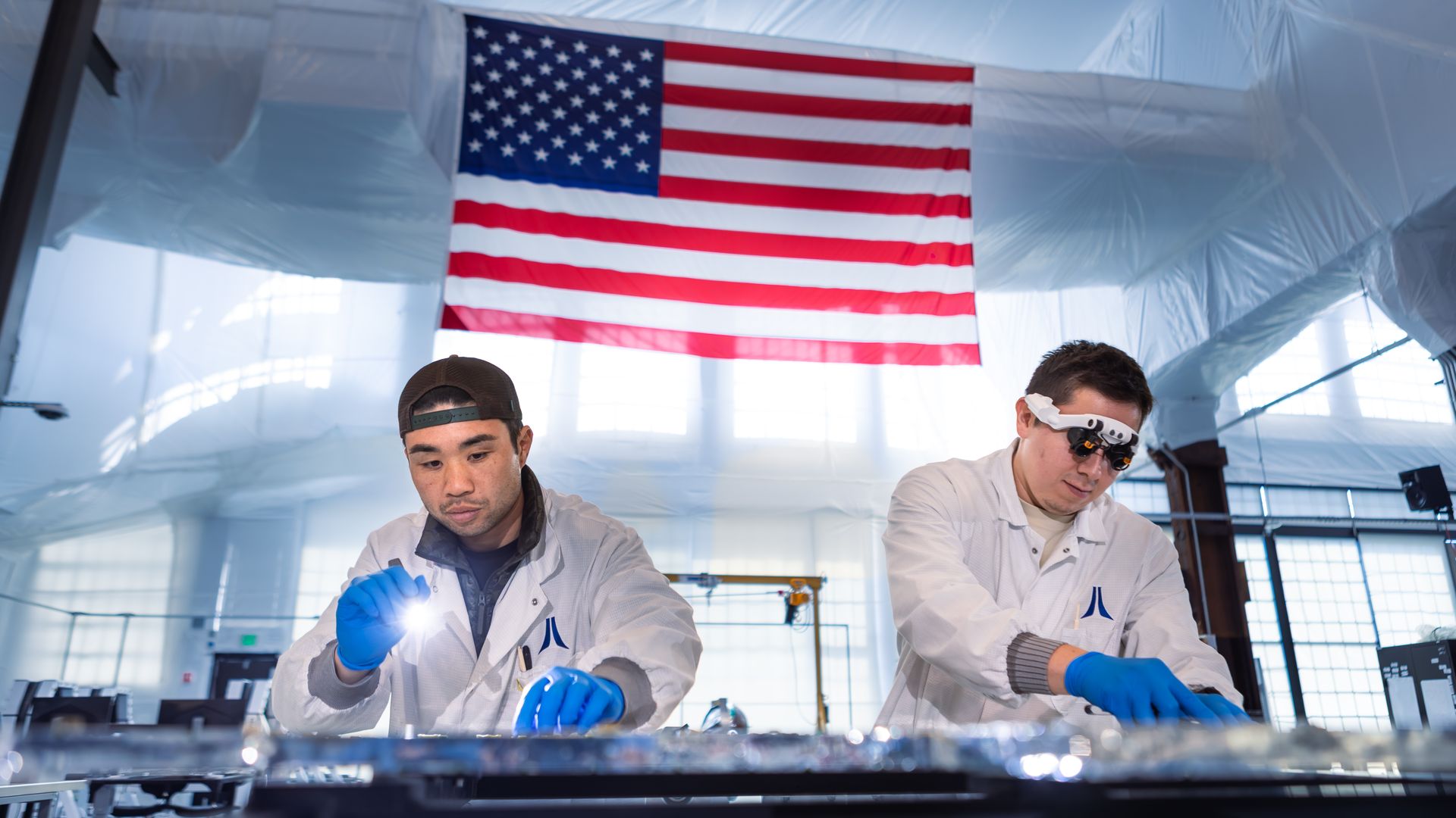 Two men in white lab coats and blue gloves work at a bench beneath a large hanging American flag in a bright, blue-toned laboratory.