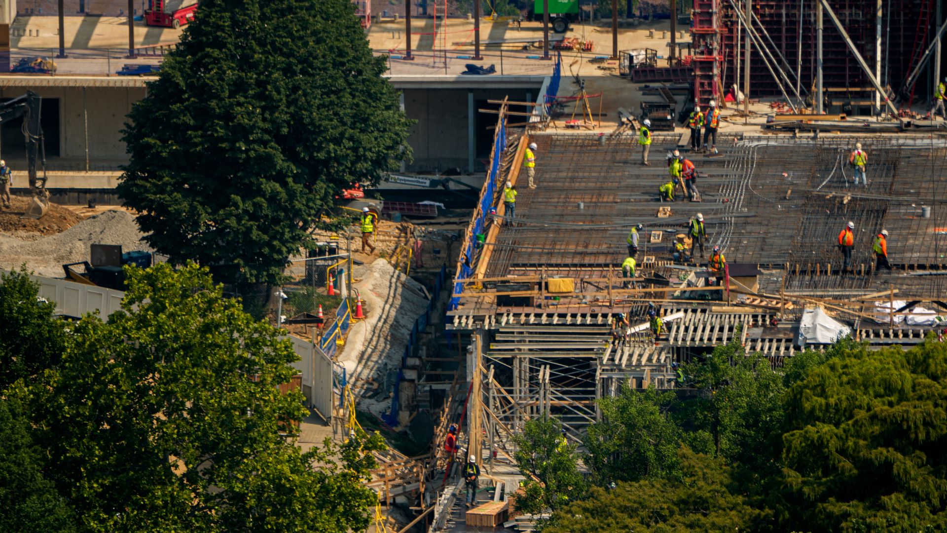 Construction workers in yellow and orange work on an in-progress project (President Trump's White House ballroom) near green trees.