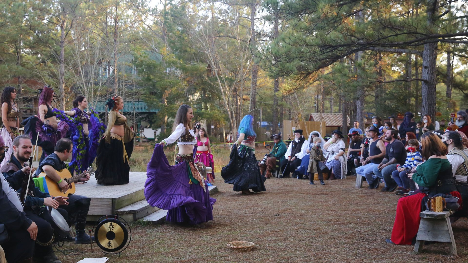 Women in Renaissance-themed outfits dance as festival visitors sit in a circle around them.