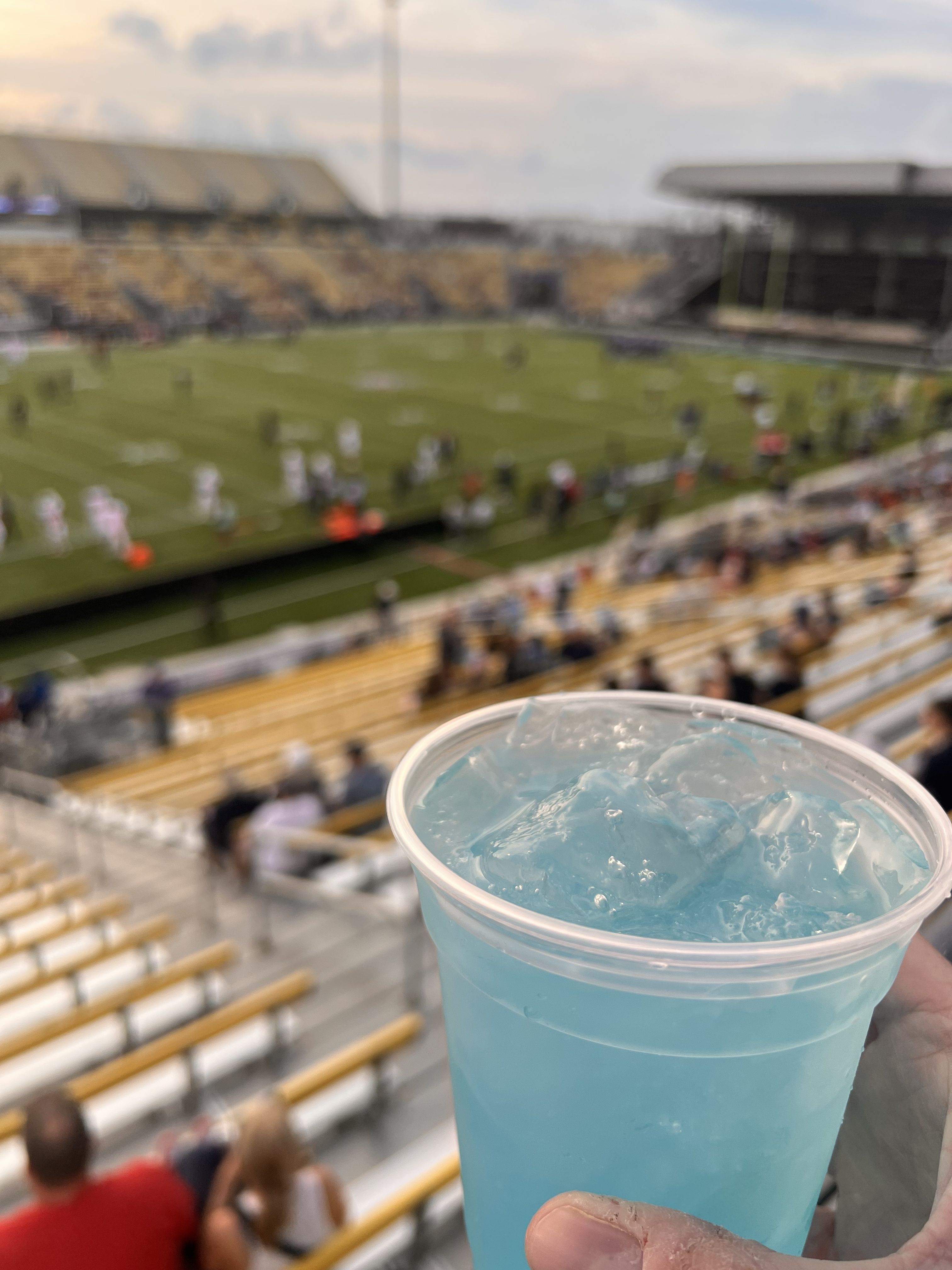 Foreground shows a clear plastic cup filled with light blue slushy ice, held by a hand. In the blurred background, a sunny football stadium with players on a green field and yellow-brown stands.