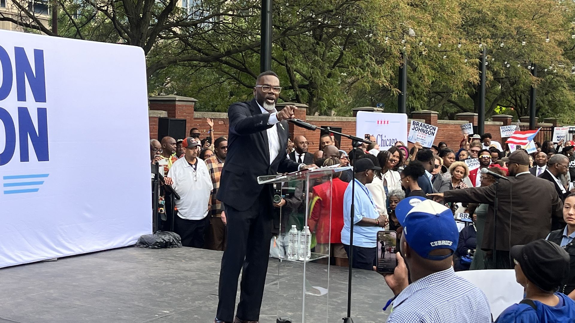 Man in a dark suit speaks at a podium during a daytime rally; a crowd holds "Brandon Johnson" signs amid trees and string lights.