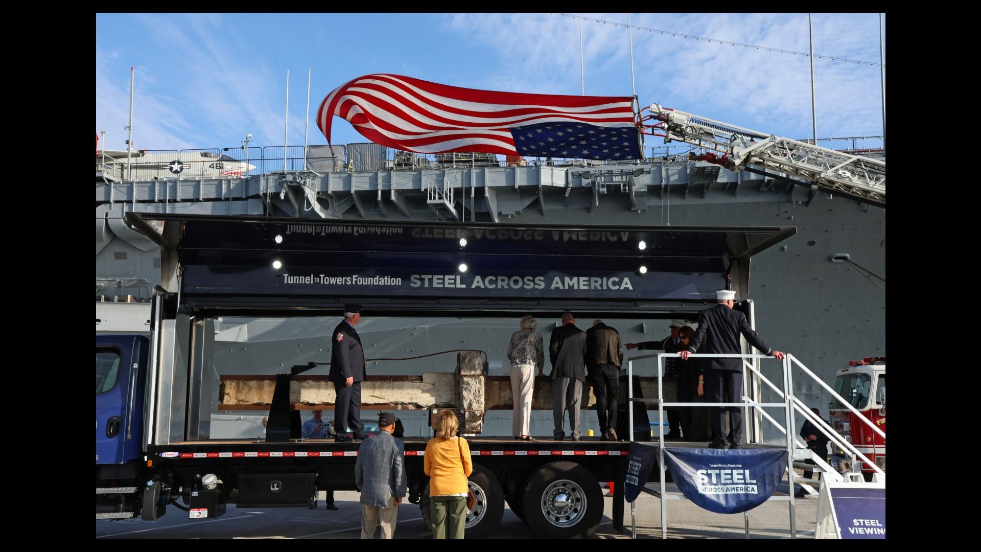 Large American flag unfurls over a gray naval ship; a banner reads "STEEL ACROSS AMERICA" from the Tunnel to Towers Foundation, as people stand on a platform beneath a blue sky.