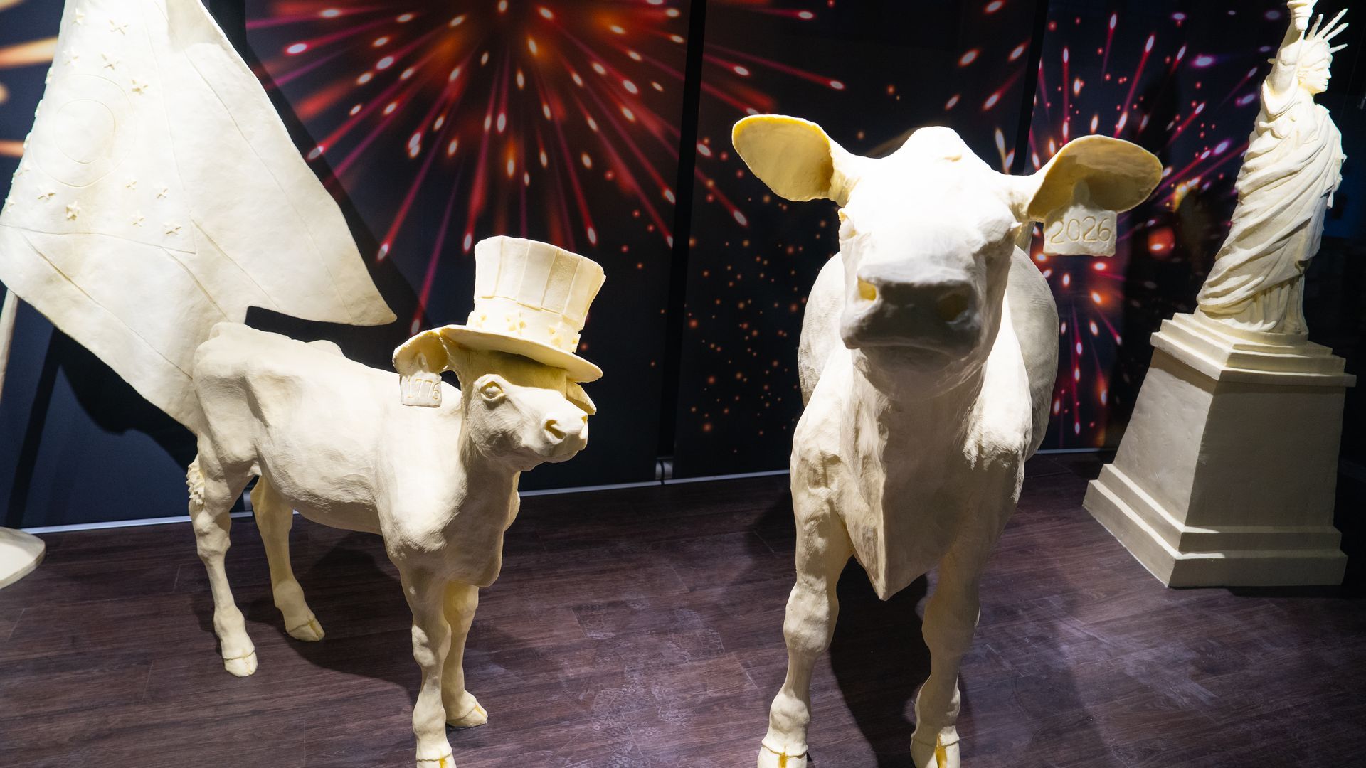 The 2026 butter cow and calf at the Ohio State Fair, in front of a backdrop of fireworks and next to a butter-sculpted Ohio flag and State of Liberty. The calf is wearing a patriotic top hat.