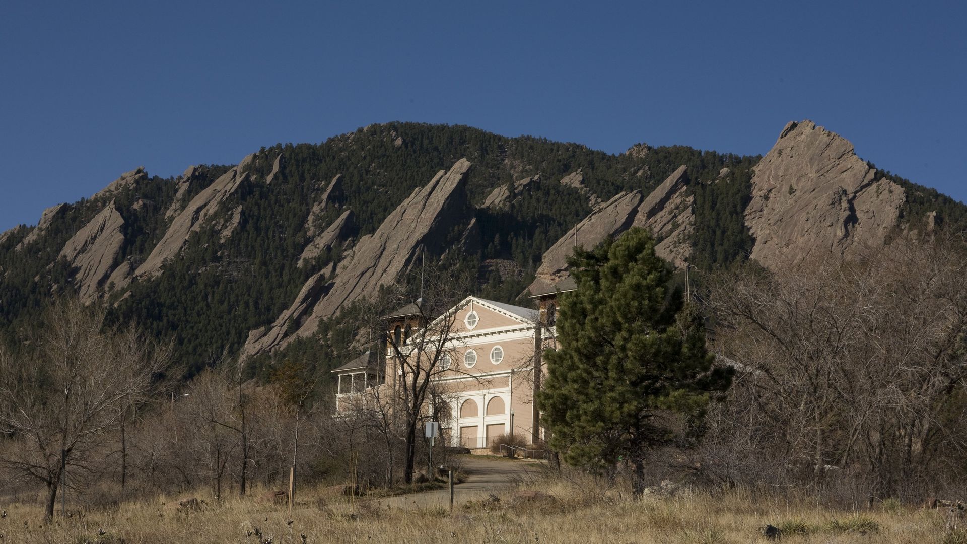 Beige building with round windows in front of rocky, forested mountain ridges under a clear blue sky, surrounded by dry grass and leafless trees.