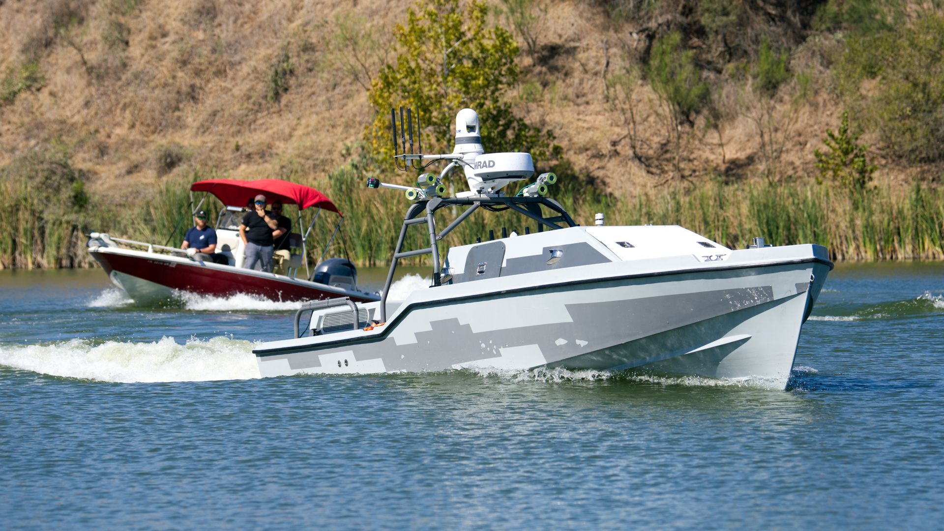 An autonomous vessel is trailed by a boat with multiple people on it. Wake splashes behind the two. In the background, brown and green brush.