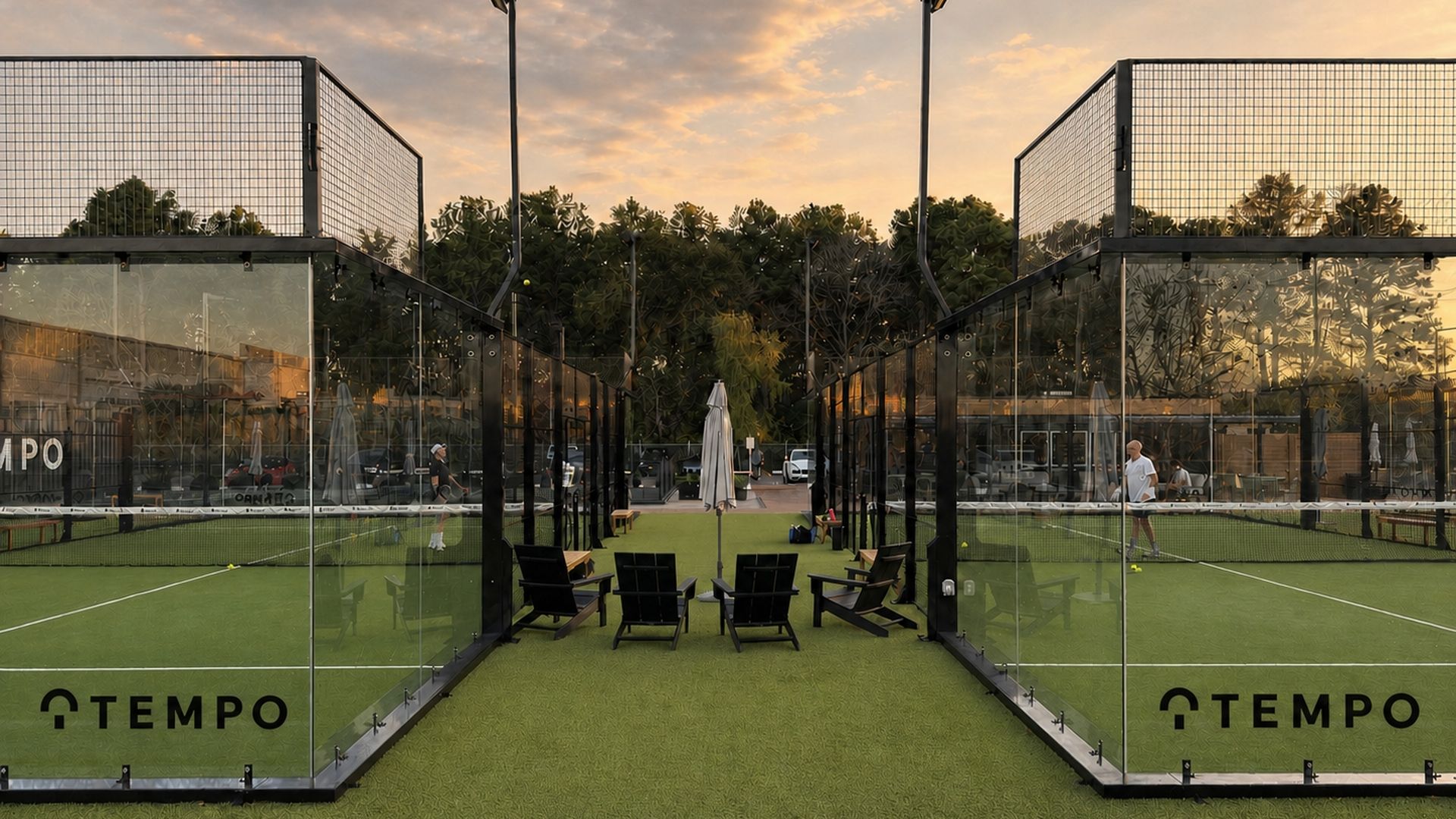 Two glass-walled padel courts with black frames flank a central seating area of chairs and umbrellas on artificial turf; sunset sky, trees in background; TEMPO branding on panels.