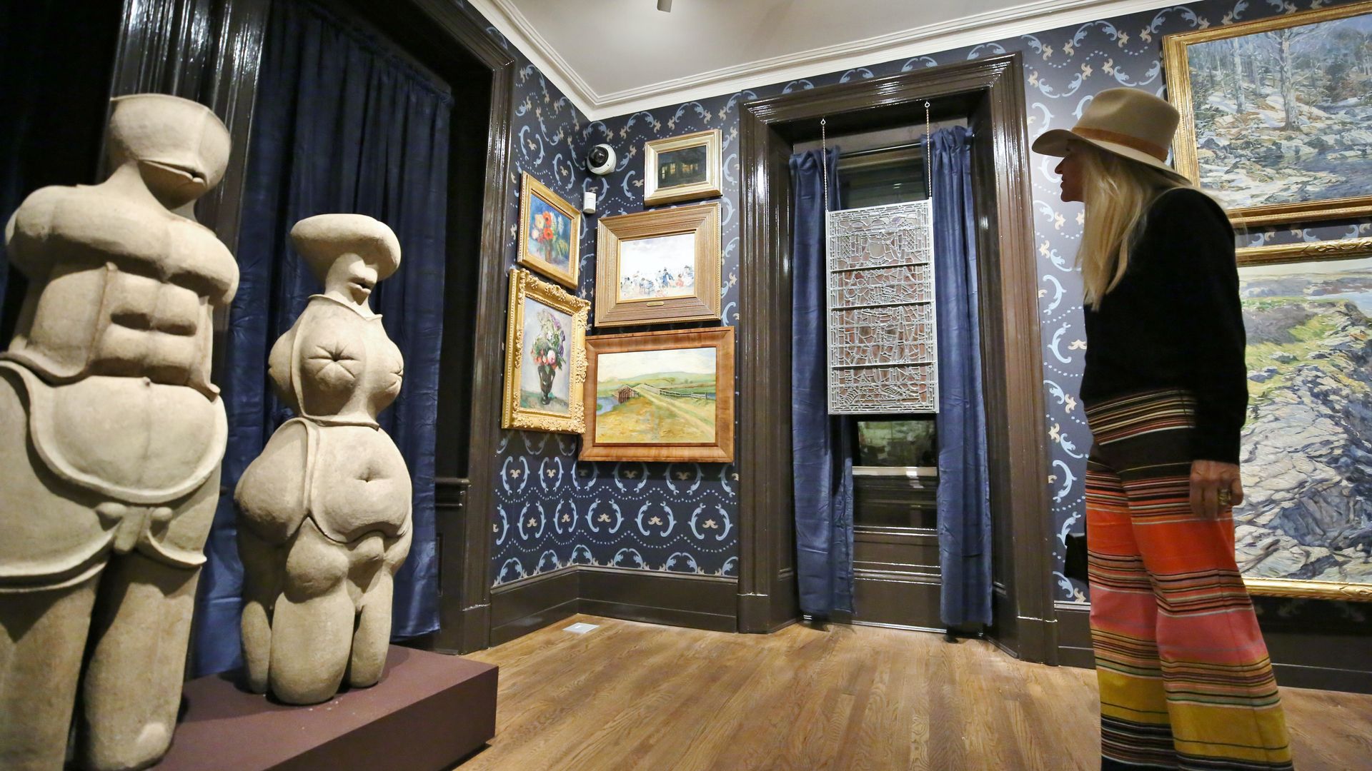 Woman in a hat and colorful striped pants views two beige abstract sculptures and several framed paintings in a blue-patterned museum room with wooden floors.