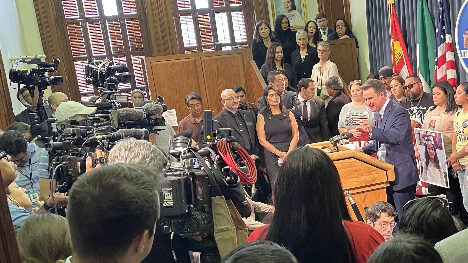 A man at a podium talks to a room filled with onlookers and tv cameras