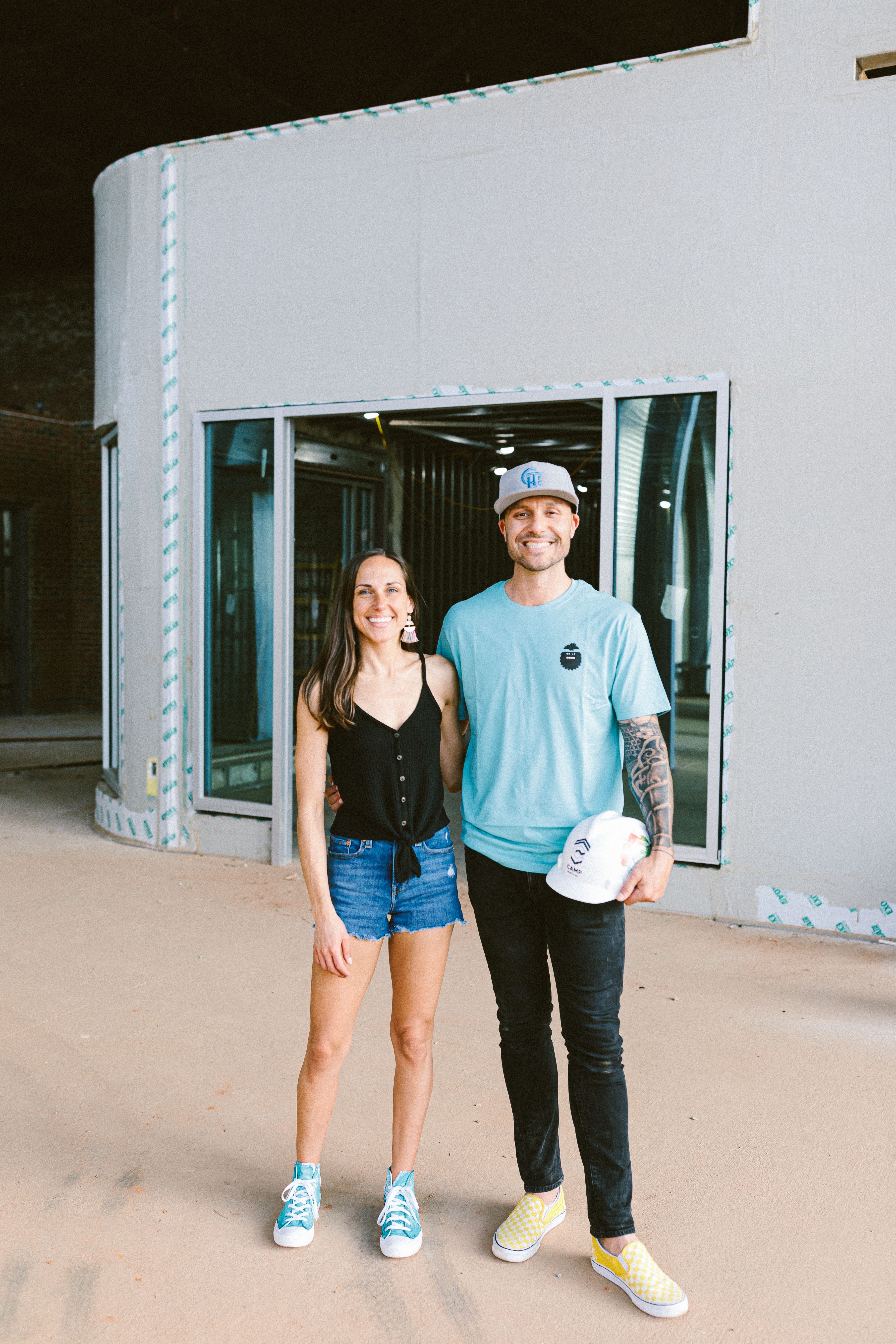 Smiling woman in black tank top, denim shorts, blue sneakers stands next to tattooed man in light blue shirt, black pants, yellow checkered shoes, holding a white helmet, indoors.