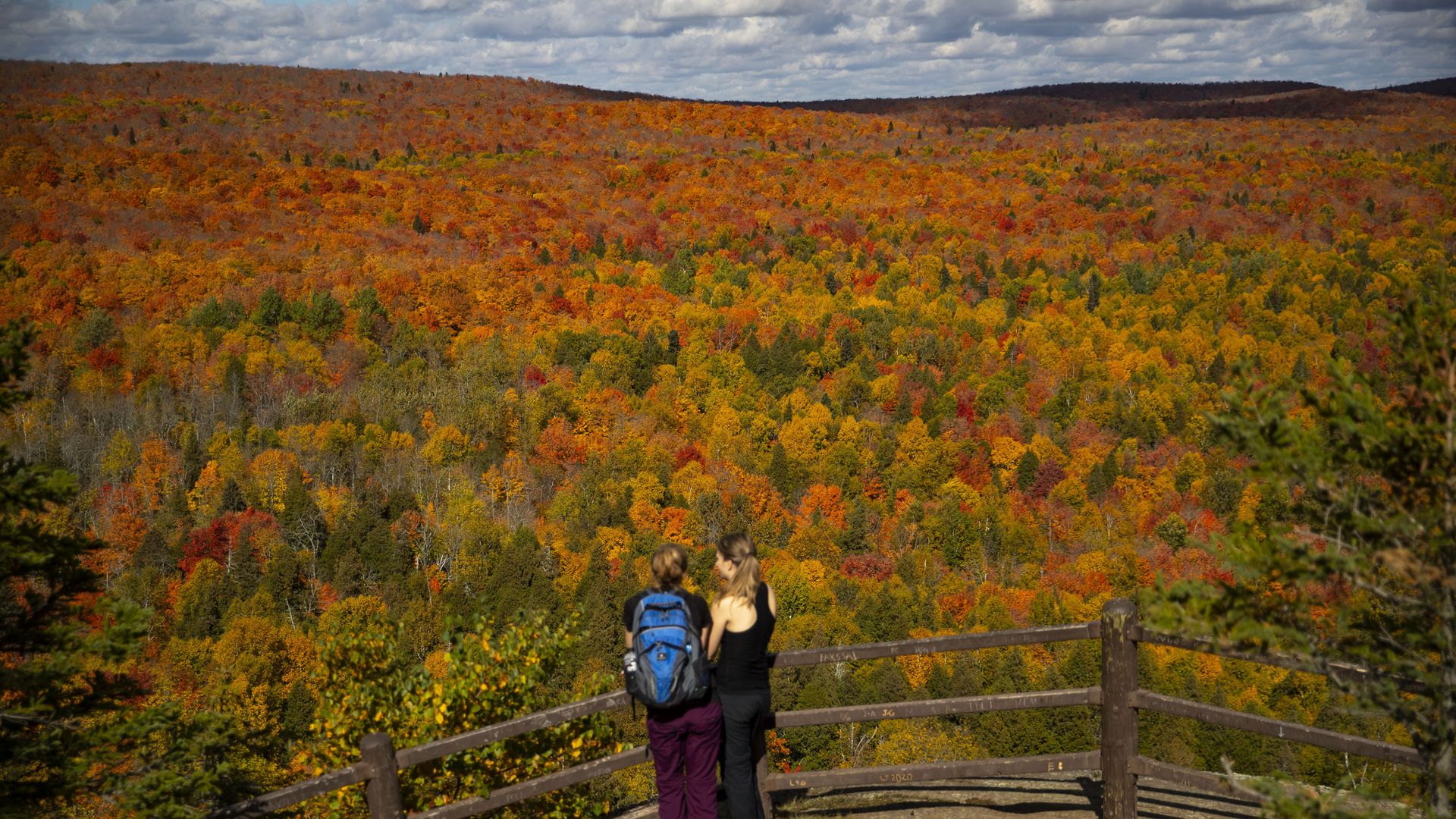 Two hikers stand at a timber fence overlooking a forest with brilliant orange, red and yellow fall foliage
