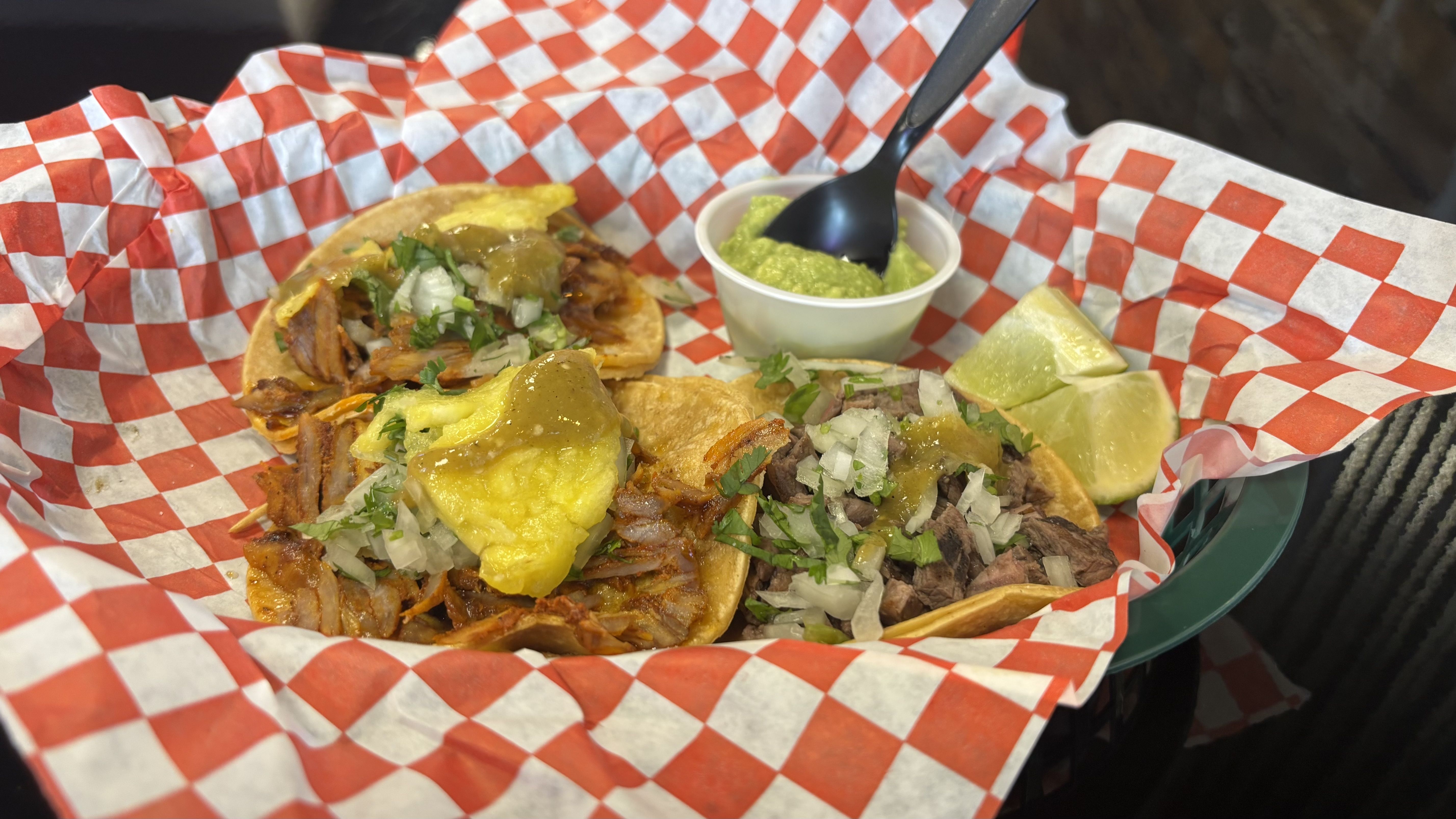 Three tacos with meat, cilantro, onions, and green salsa on yellow corn tortillas served on red-and-white checkered paper with lime wedges and green guacamole in a small cup.