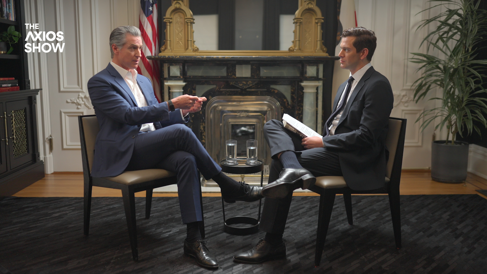 Two suited men sit across from each other in a formal room with a marble fireplace, a small table, and two glasses of water; one gestures while the other holds a newspaper.