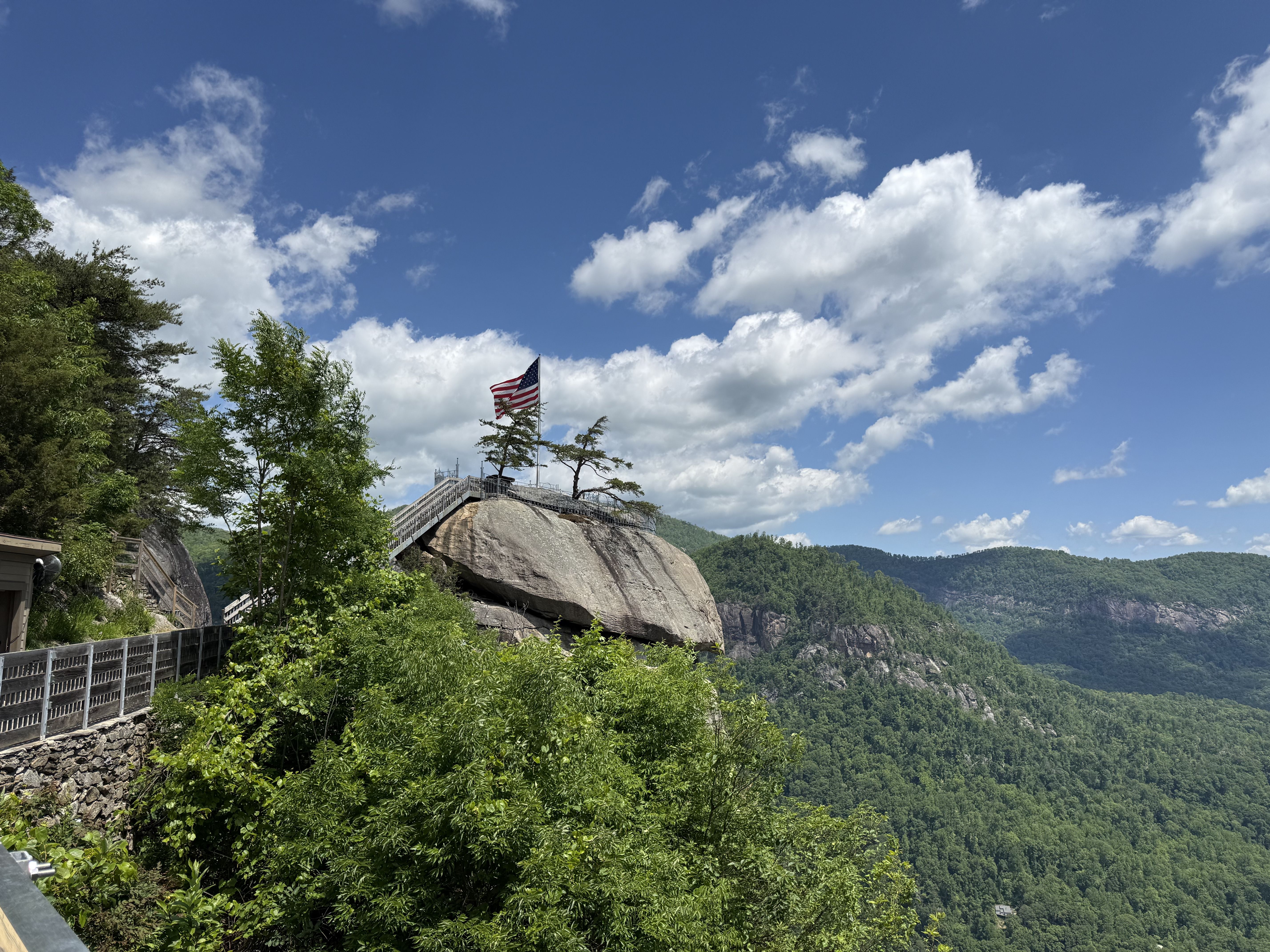 Bright blue sky with white clouds above a rocky overlook. A metal staircase and railing lead to a flat summit where an American flag waves, surrounded by green trees and distant mountains.