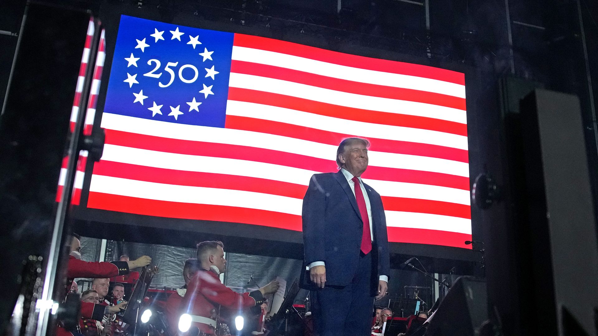 Large display of the American flag with a blue canton and a 250-star ring; a man in a dark suit and red tie stands on stage, while a band and red-clad audience are visible below.