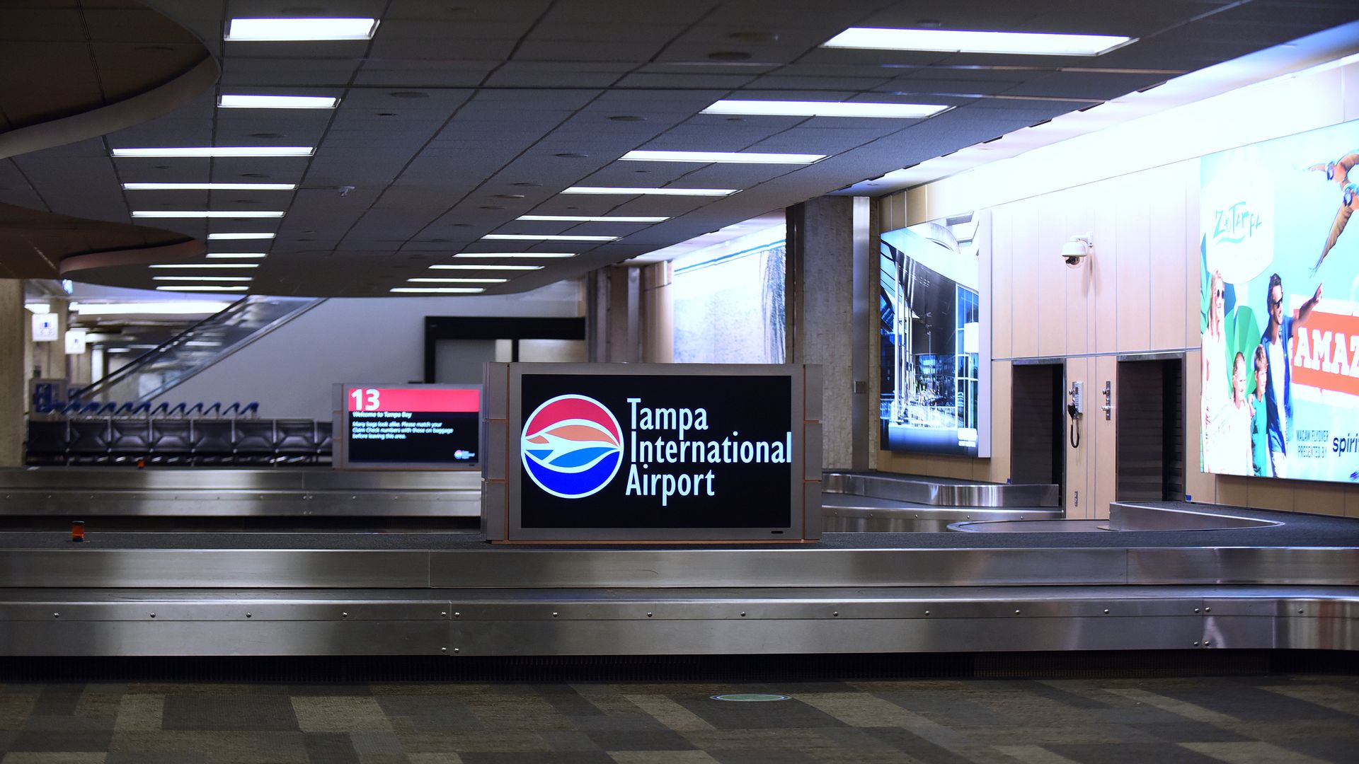 Empty baggage claim area at Tampa International Airport with a sign displaying the airport's name and logo in red, blue, and white. Advertisement panels are visible on the right wall.