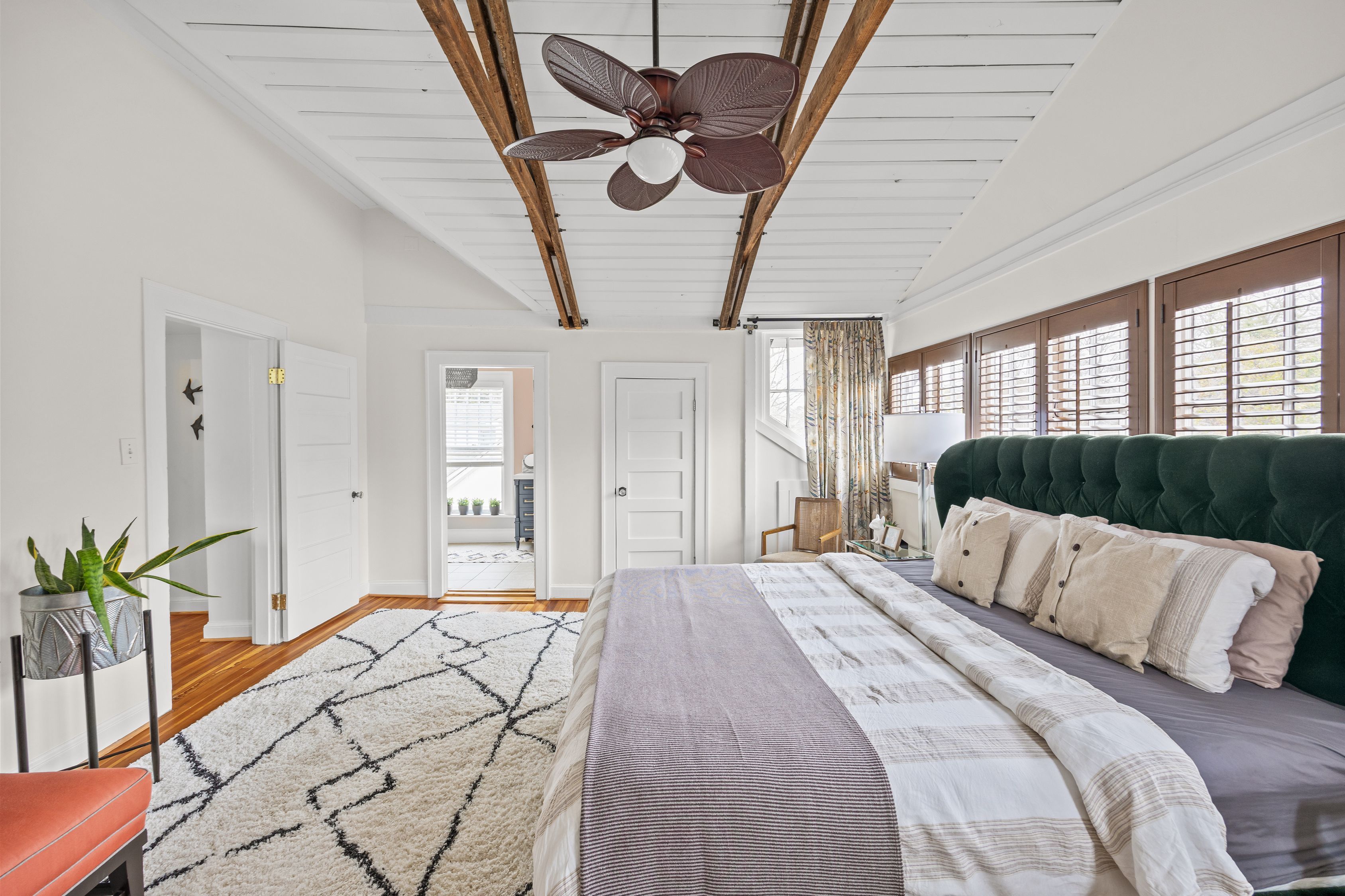 Bright bedroom with white walls, wooden beams, dark green tufted headboard, beige and white bedding, patterned rug, wooden window shutters, ceiling fan, and potted plant on stand.