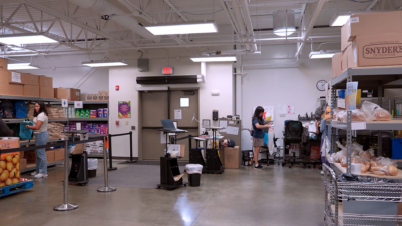 Warehouse-style grocery with bright lights. Left: a woman browsing shelves of bags and boxes. Center: checkout area with laptop. Right: shelves stacked with bread and groceries.