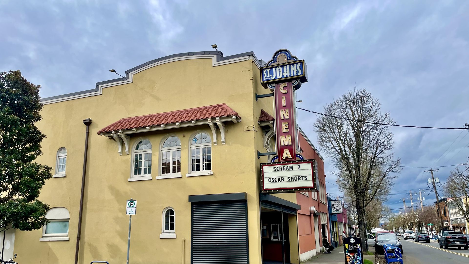 Exterior of St. Johns Cinema with yellow walls, red tiled roof accents, and a vertical sign showing "SCREAM 7" and "OSCAR SHORTS" under a cloudy sky on a street with parked cars.