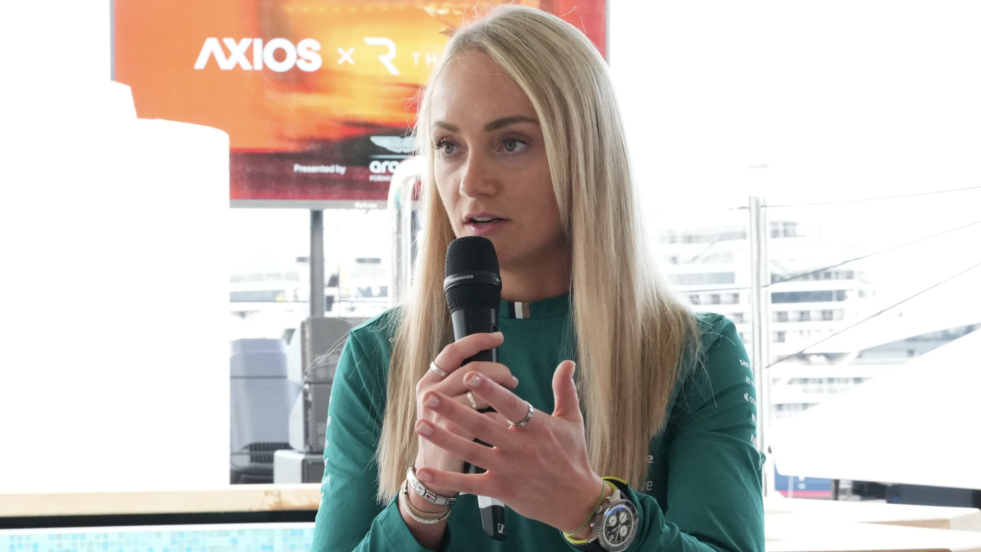 Blonde woman in a green top speaks into a handheld microphone at an event; a bright red-orange banner appears behind her.