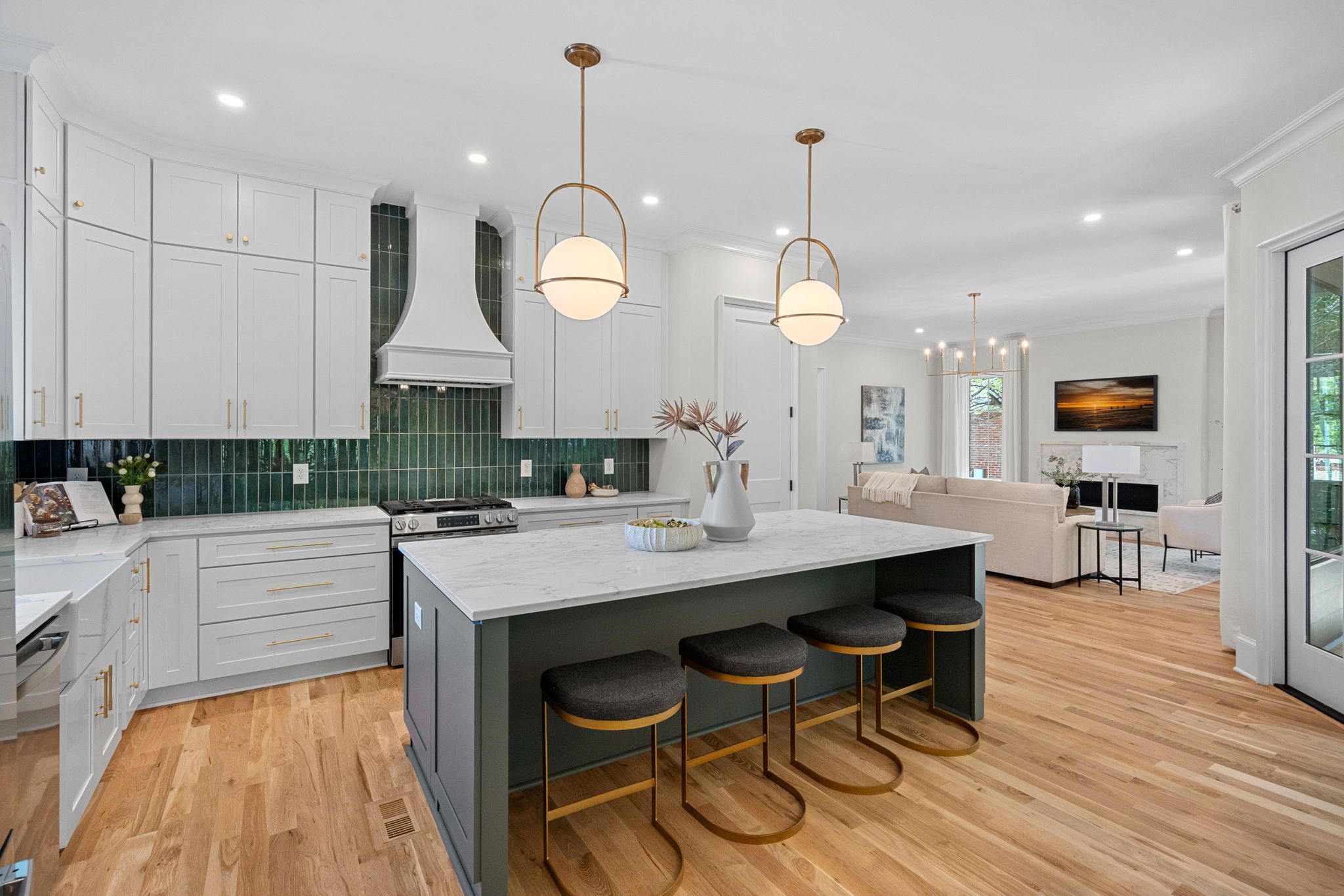 Bright kitchen with white cabinets, dark green tile backsplash, marble countertops, and a dark island with three gold-framed stools. Gold pendant lights; living area with sofa and fireplace beyond.