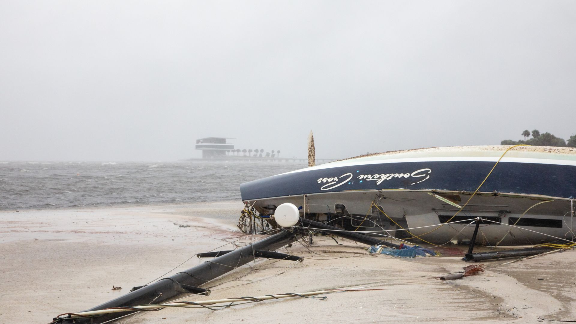 A sailboat grounded on a sandy shore and turned on its side with gray water, a gray sky and a pier in the background.