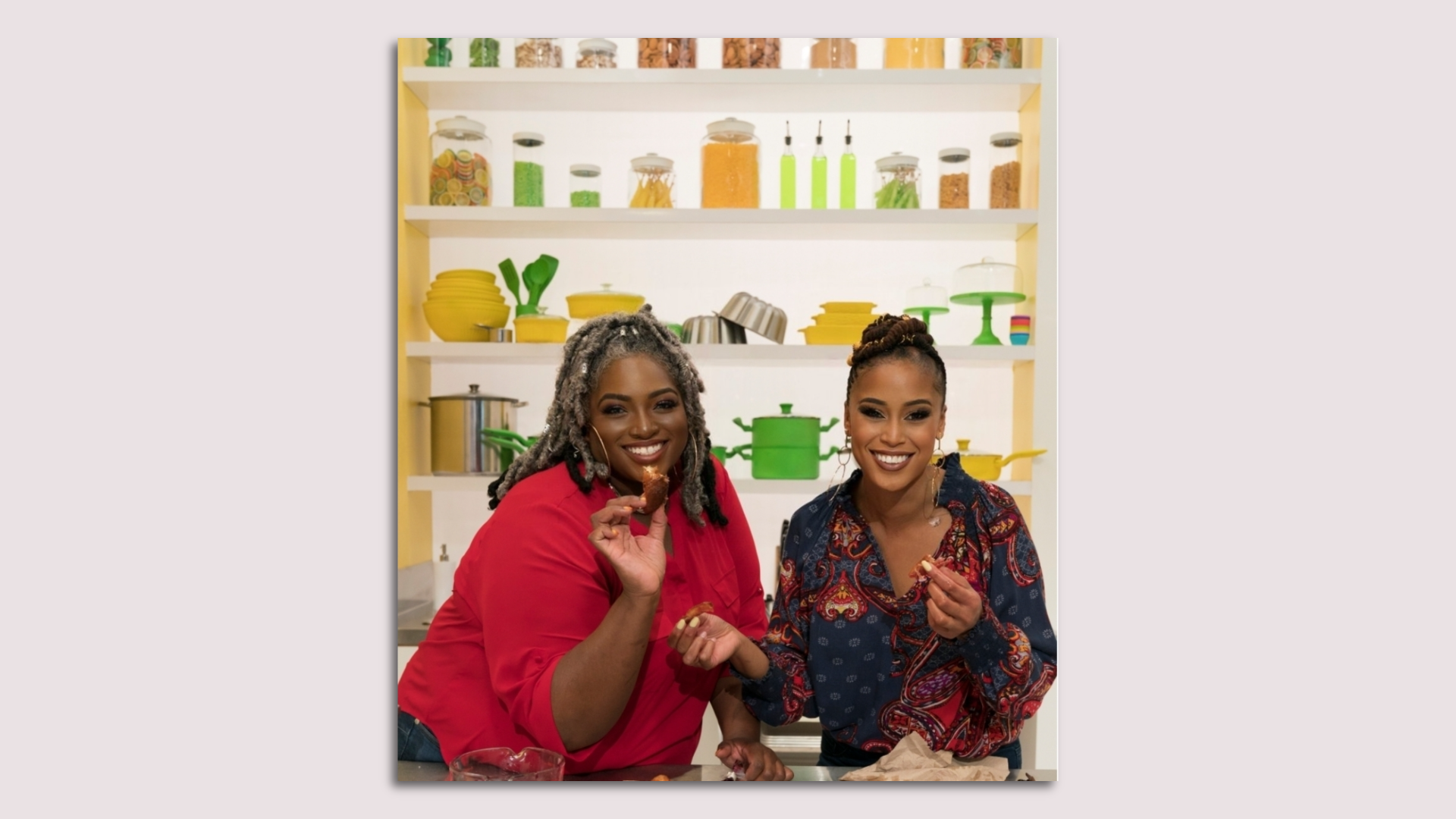 Two women hold beignets against a pink background