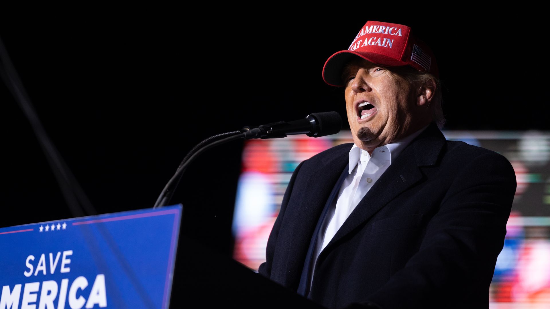 Photo of Donald Trump speaking from a podium with a MAGA hat