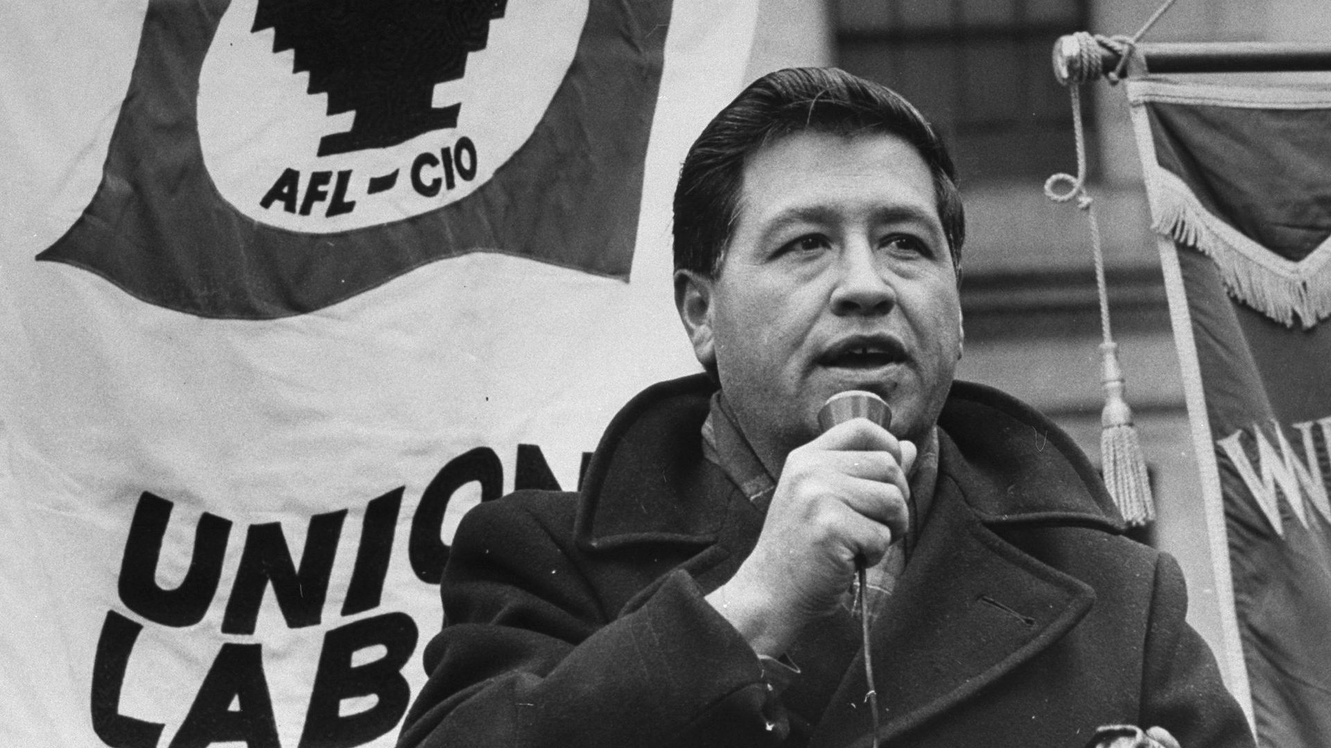 Black-and-white photo of a man speaking into a microphone at a rally. A large flag reads 'FARMWORKERS AFL-CIO' with a stylized emblem; another banner shows 'UNION LABOR' in the background.