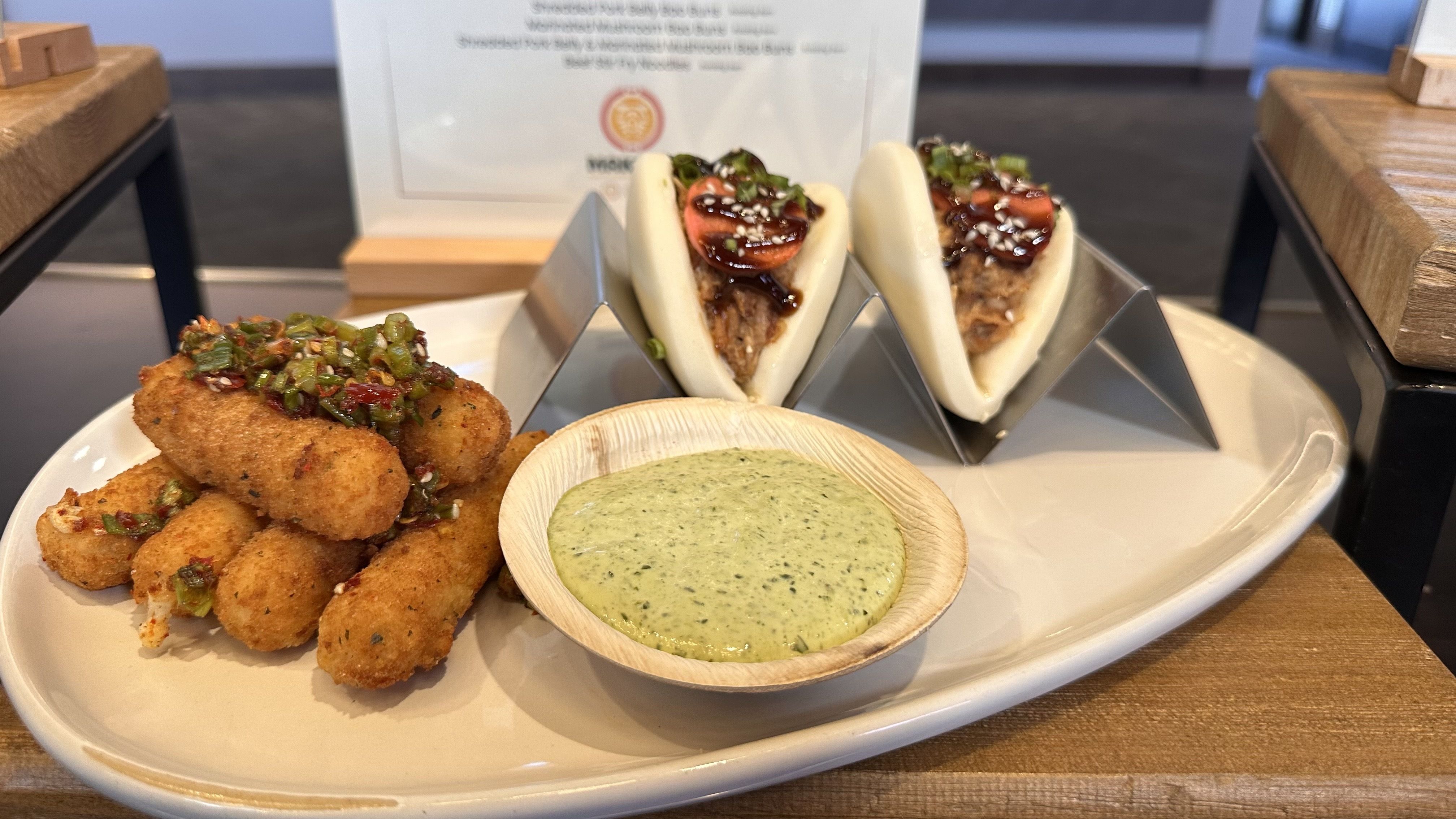 Plate of golden fried croquettes topped with chopped green peppers, two steamed bao buns with meat and dark glaze with sesame seeds, plus a pale green herb dip in a wooden bowl; wooden table.