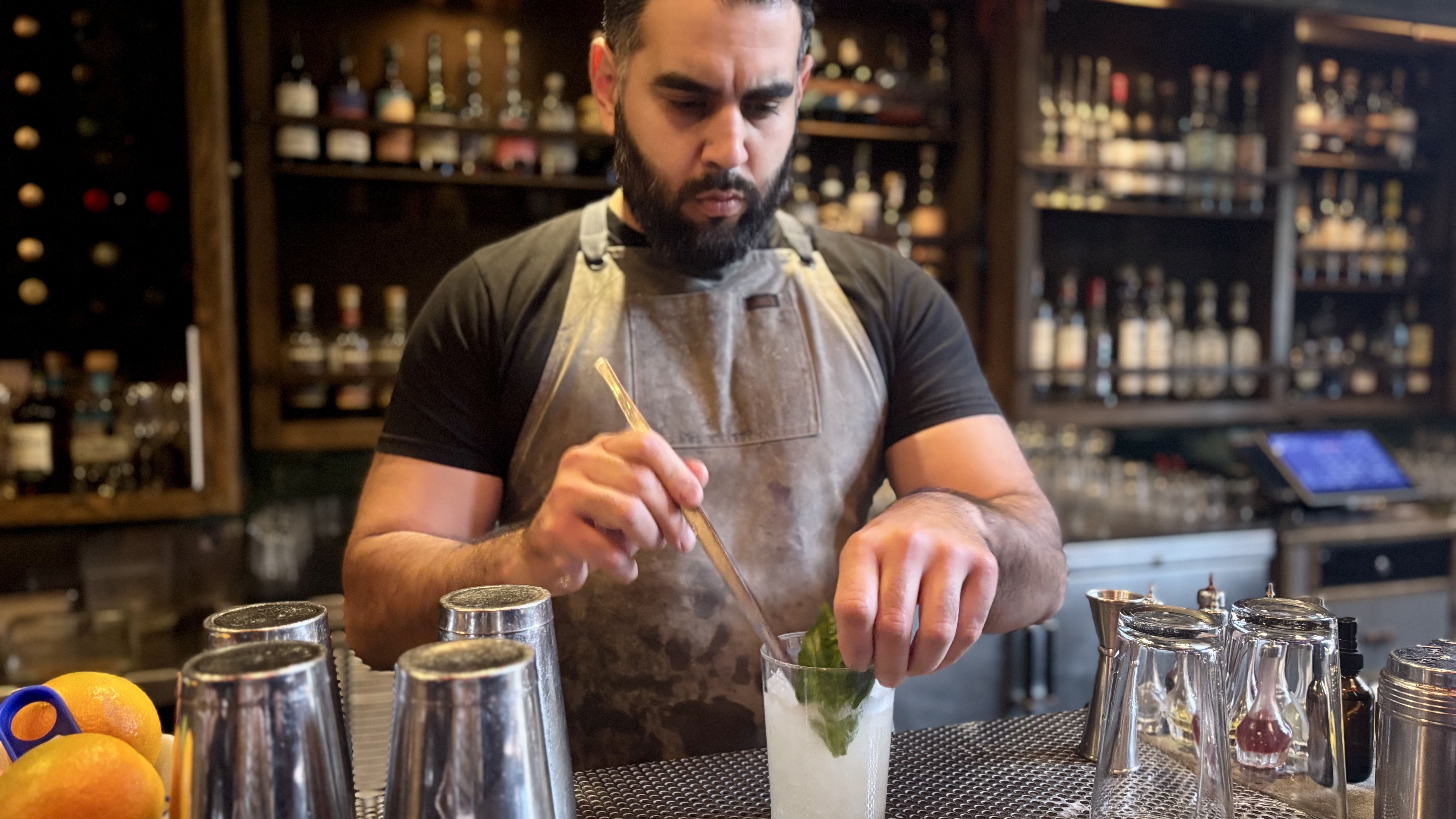 Bartender with a beard wearing a black shirt and apron stirs a cocktail with a leaf garnish at a bar with shelves of liquor bottles in the background.