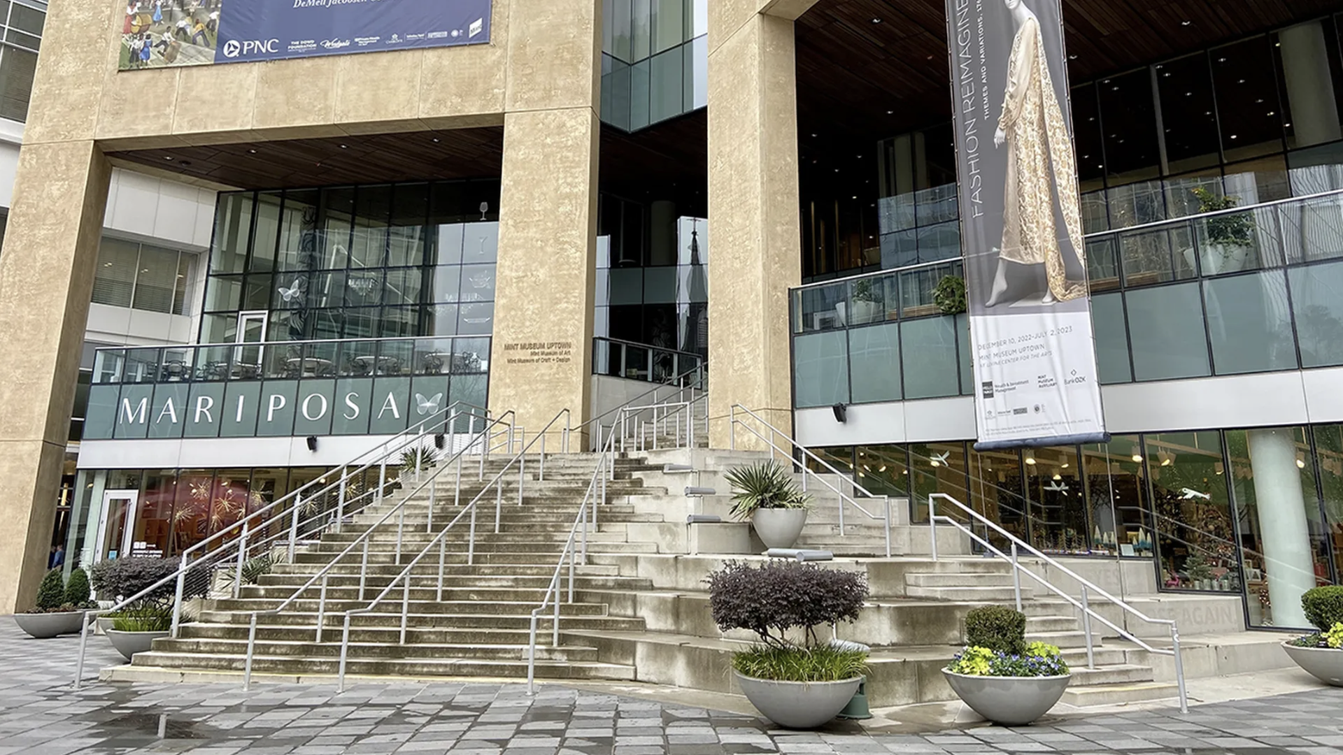 Large stone staircase with handrails leading up to a glass building entrance labeled "MARIPOSA." Banner on right shows a fashion exhibit with a model in a long ornate dress. Planters with greenery.