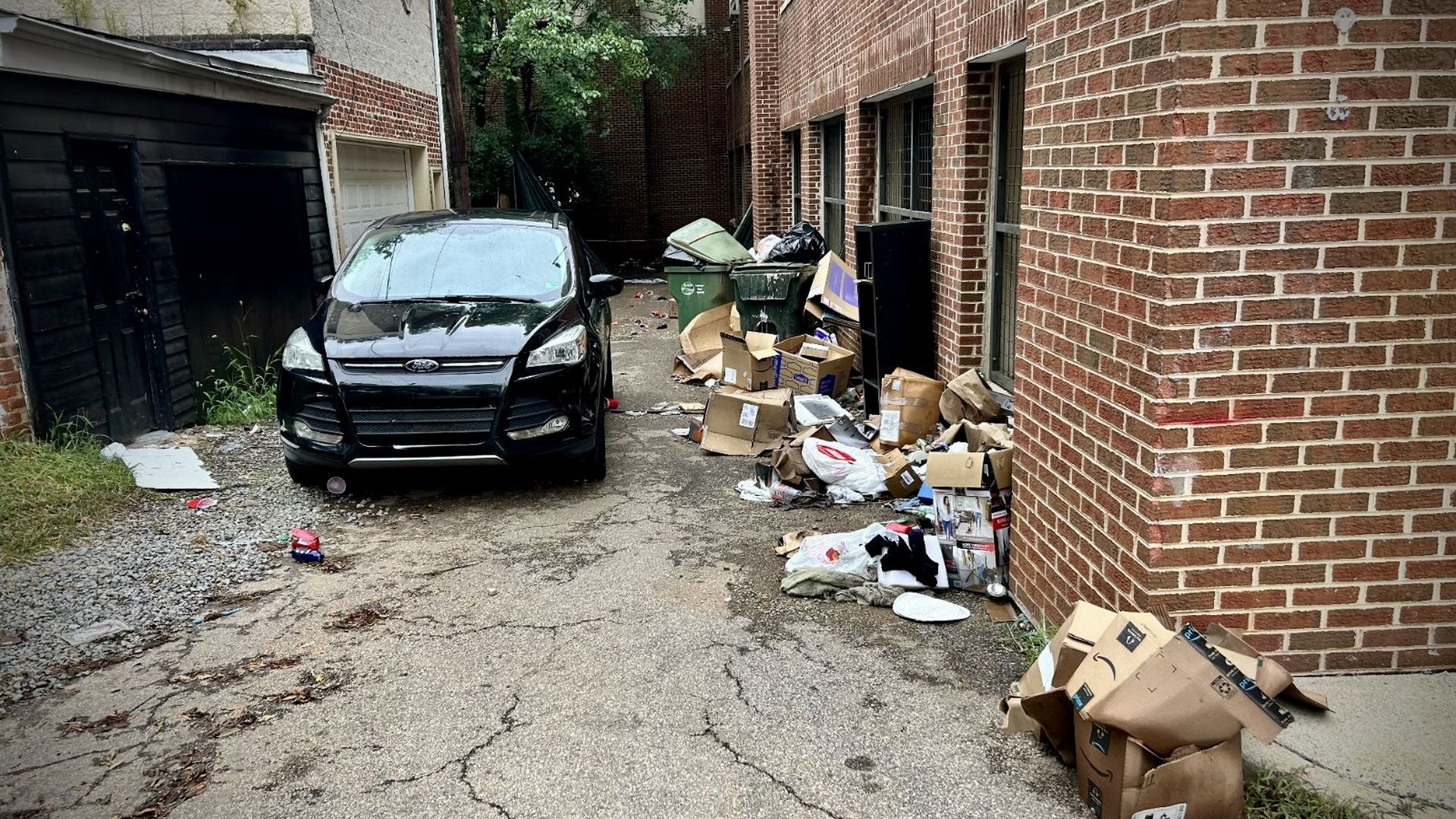 A narrow alley between brick buildings; a black Ford SUV is parked on cracked pavement. Piles of cardboard boxes and trash bags sit by green and black dumpsters, with a shed and trees to the left.