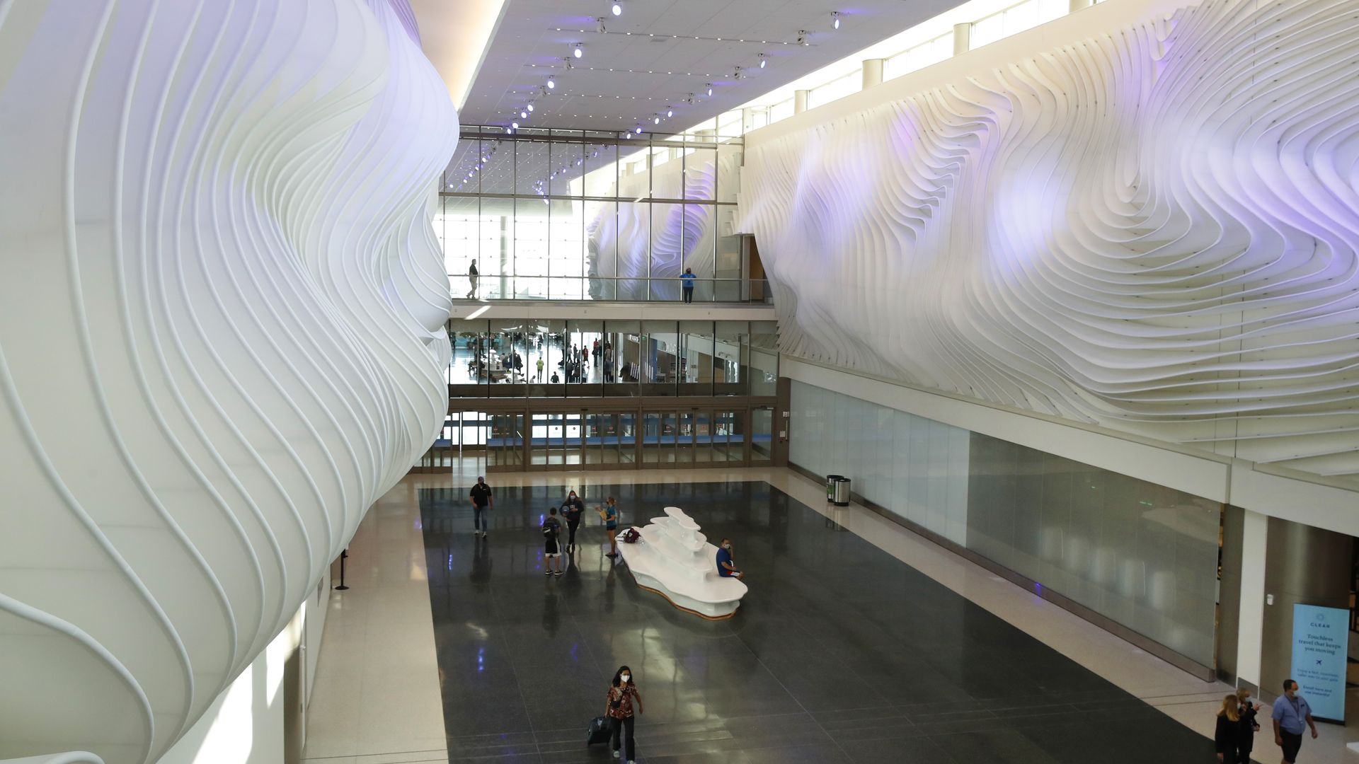 Inside a modern, airy atrium with white, wave-like sculptural walls, soft purple lighting, glass balconies, and people walking on a dark, glossy floor.