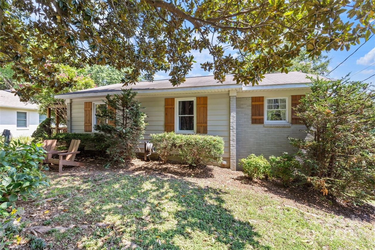 Single-story beige house with wooden shutters and shrubs in front, two wooden chairs on left lawn area beneath leafy tree branches on a sunny day.
