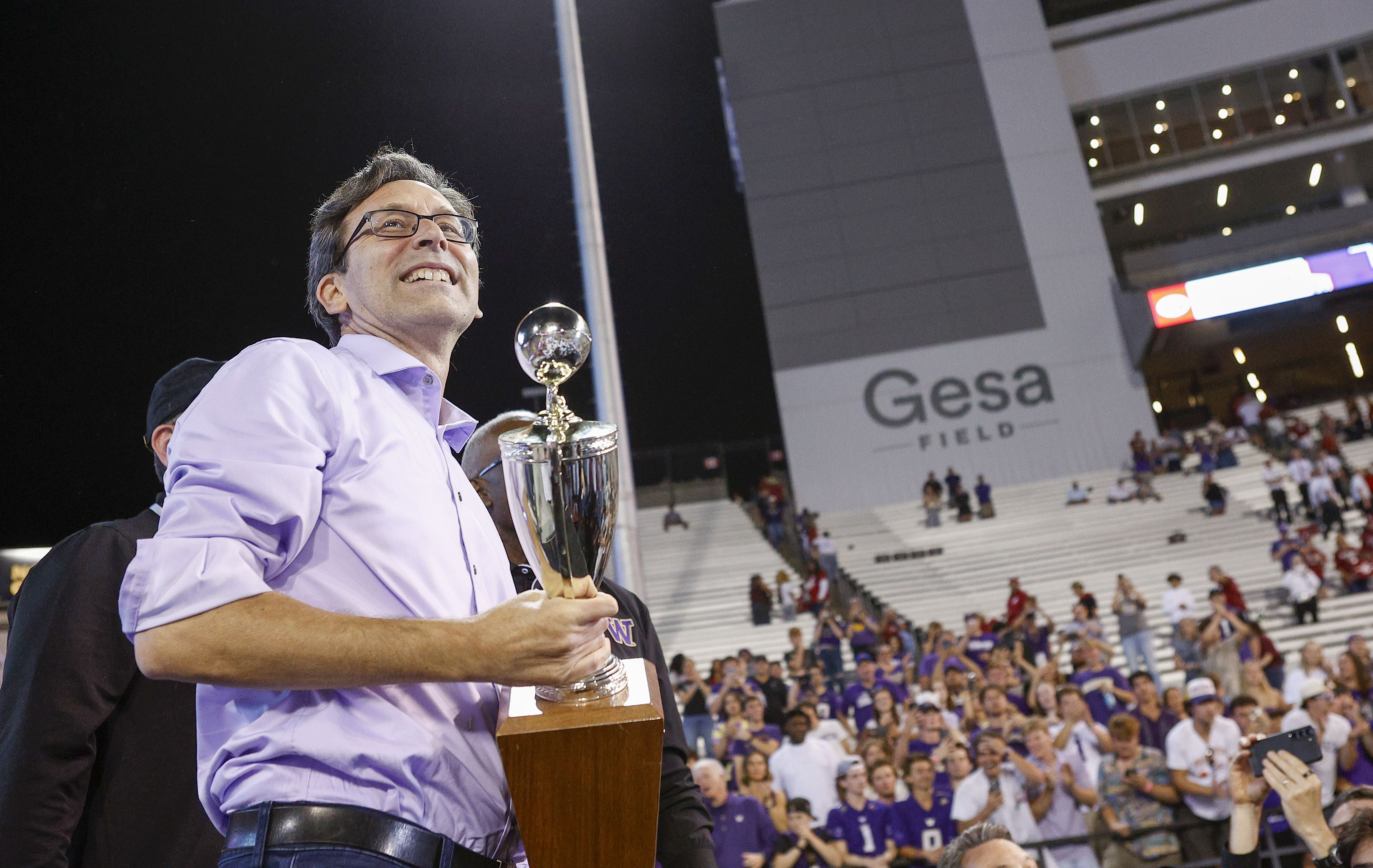 Bob Ferguson wears a light purple button up shirt rolled up to his elbows as he holds the Apple Cup, smiling, with a crowd in the stadium seats behind him. at night time. 