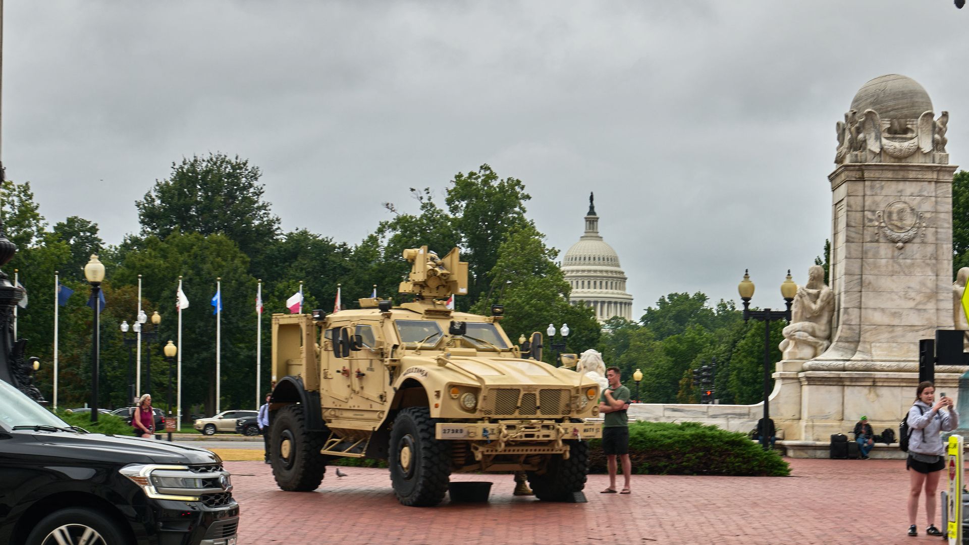 Beige military armored vehicle parked on a brick plaza with the U.S. Capitol visible in the background and people nearby, some taking photos, under a cloudy sky.