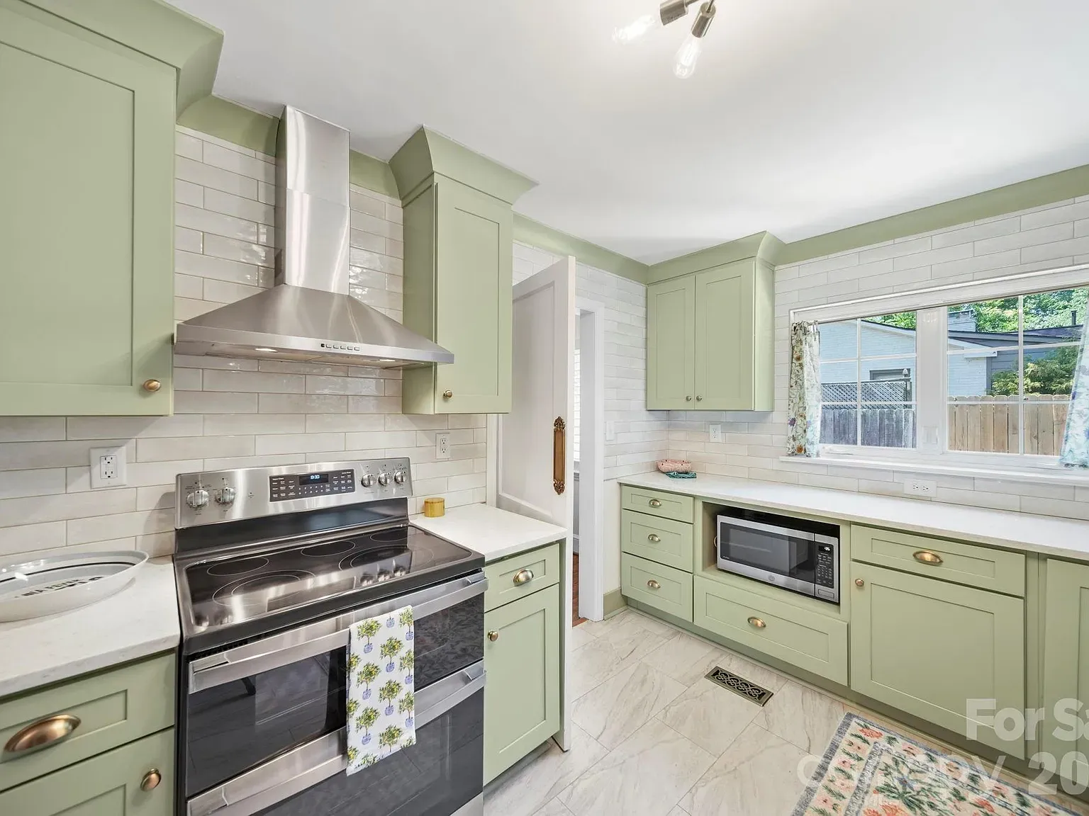 Bright kitchen with pale green cabinets, white subway-tile backsplash, stainless steel range hood and stove. White countertops, a microwave, and a large window with floral curtains; light wood floor.