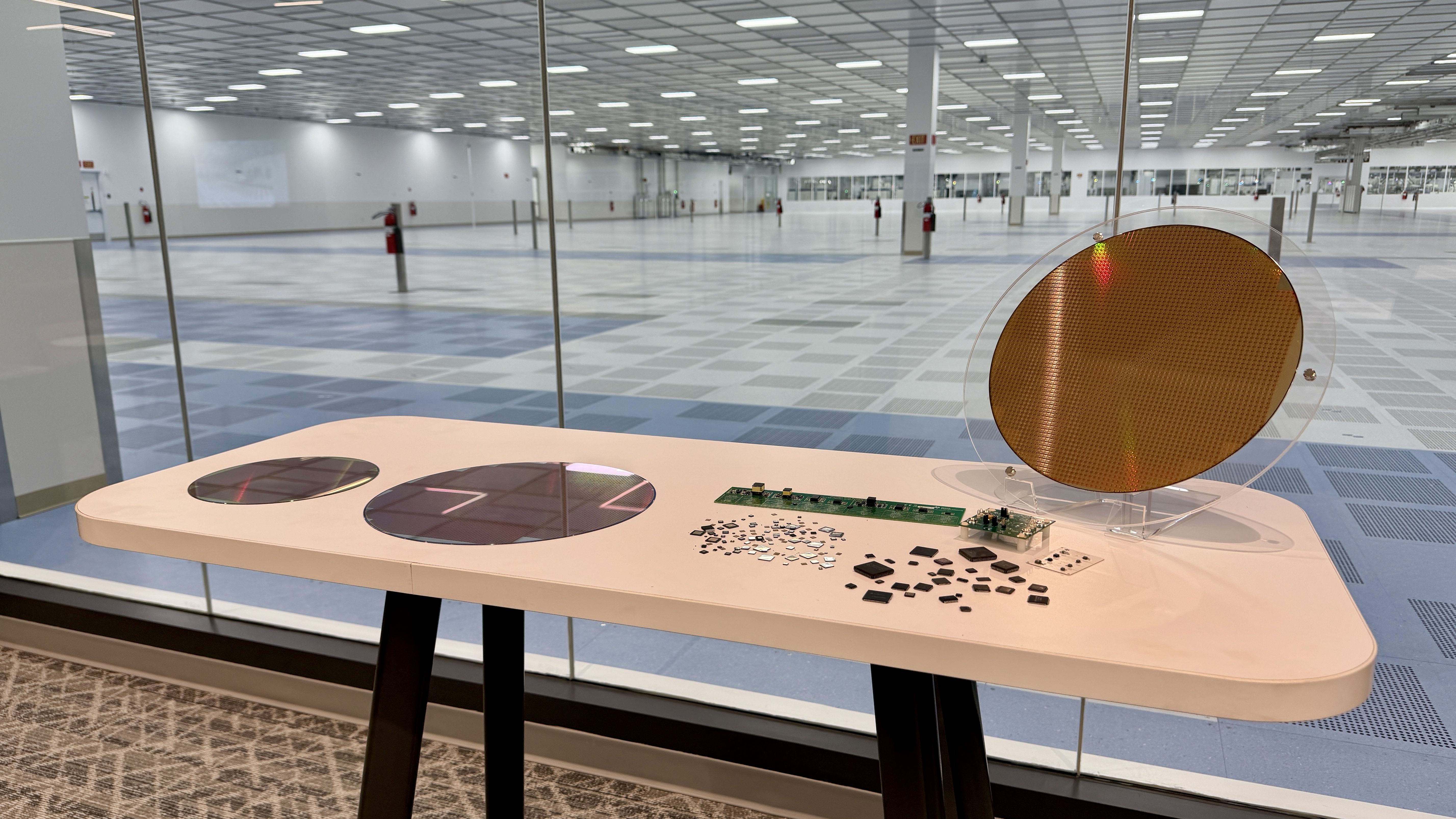 Bright, empty exhibition hall seen through glass walls; in the foreground a white display table features a large orange circular disk and scattered electronic components.