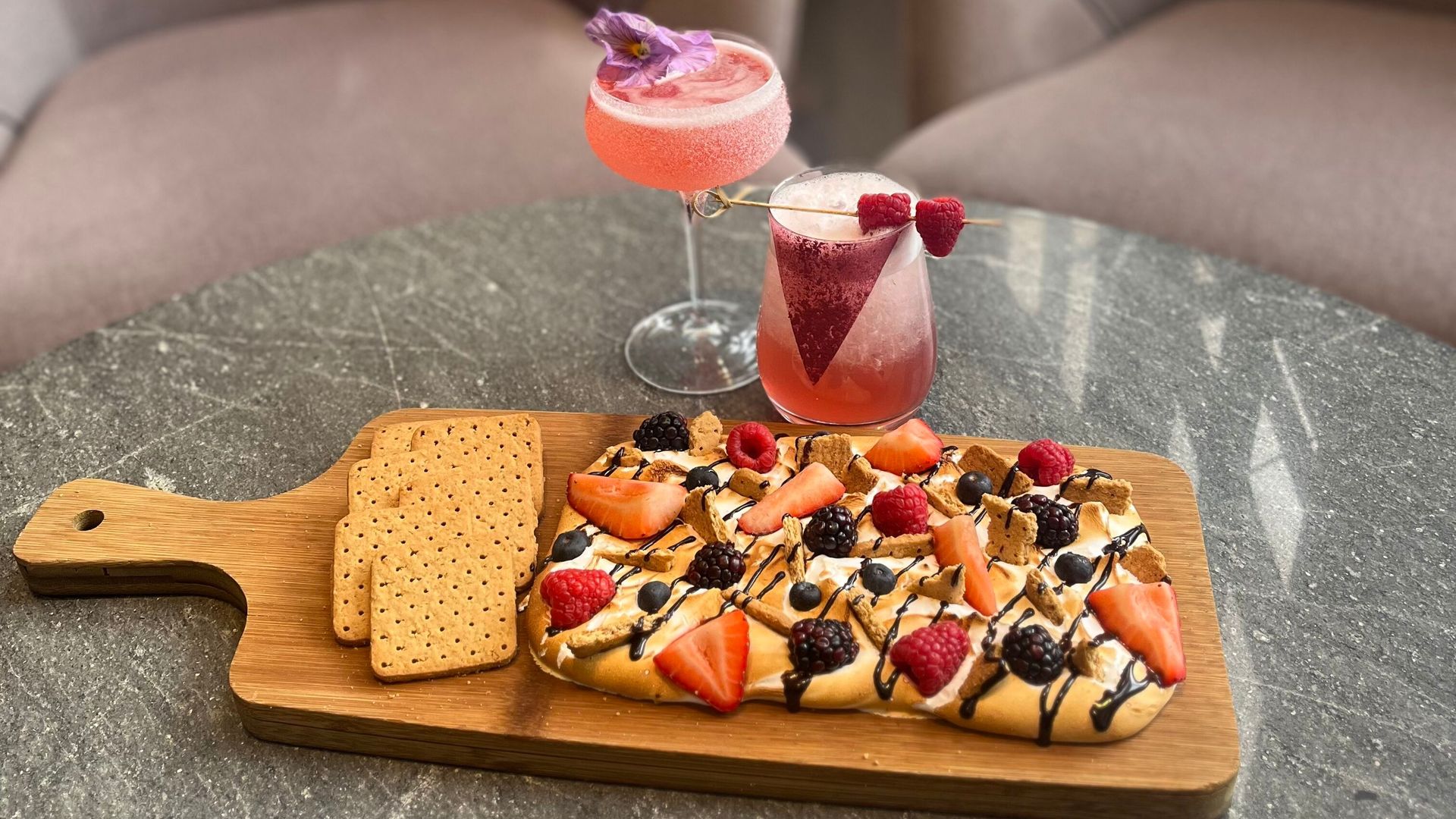A s'mores board with two pink cocktails besides it.