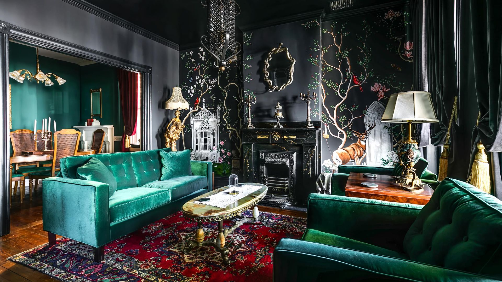 A dark room with dark green velvet sofas, black walls, older spooky looking decor, black drapes and a chain-like chandelier.