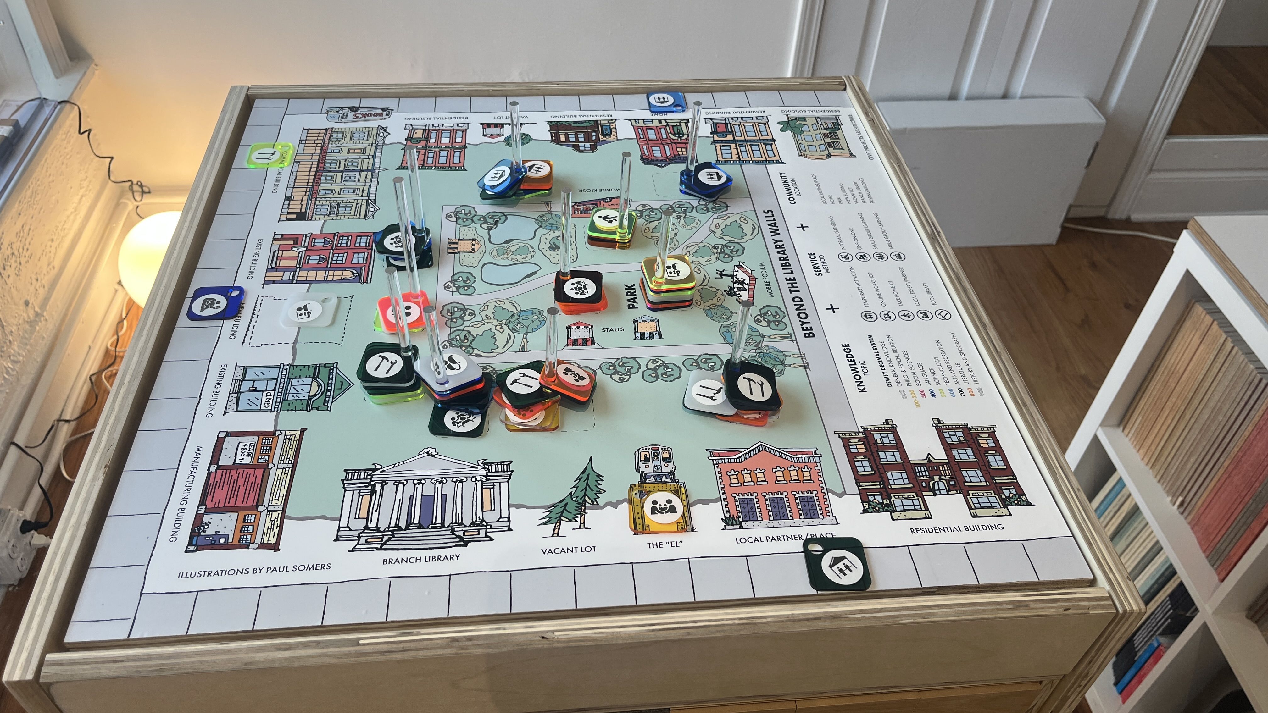 A colorful board game titled "Beyond the Library Walls" on a wooden table, featuring illustrated buildings, parks, and game pieces with icons on clear stands in a cozy room with bookshelves.