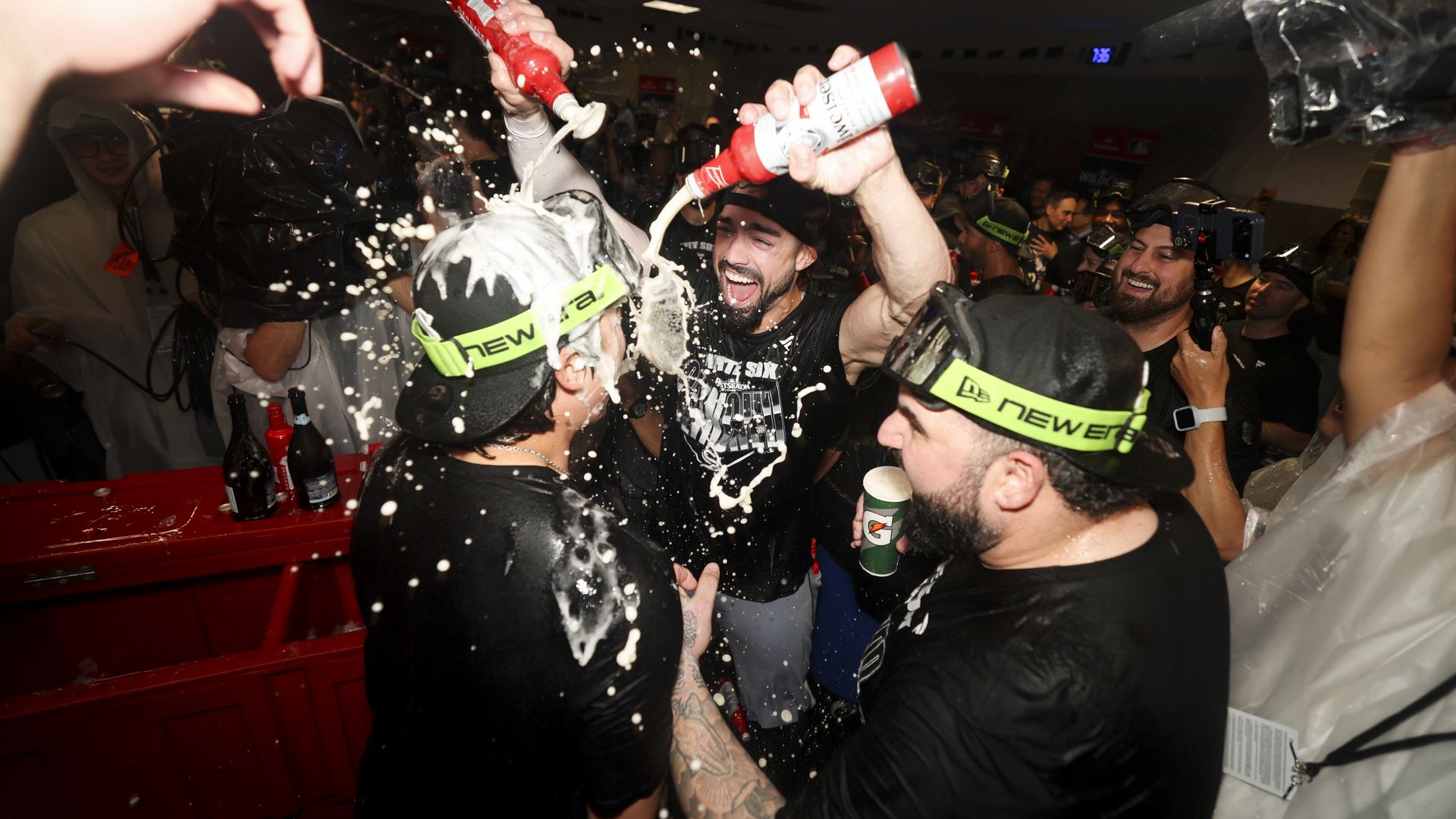 An indoor party with a crowd cheering as foam sprays from red bottles; people in black shirts and neon headbands raise drinks amid a chaotic, festive scene.