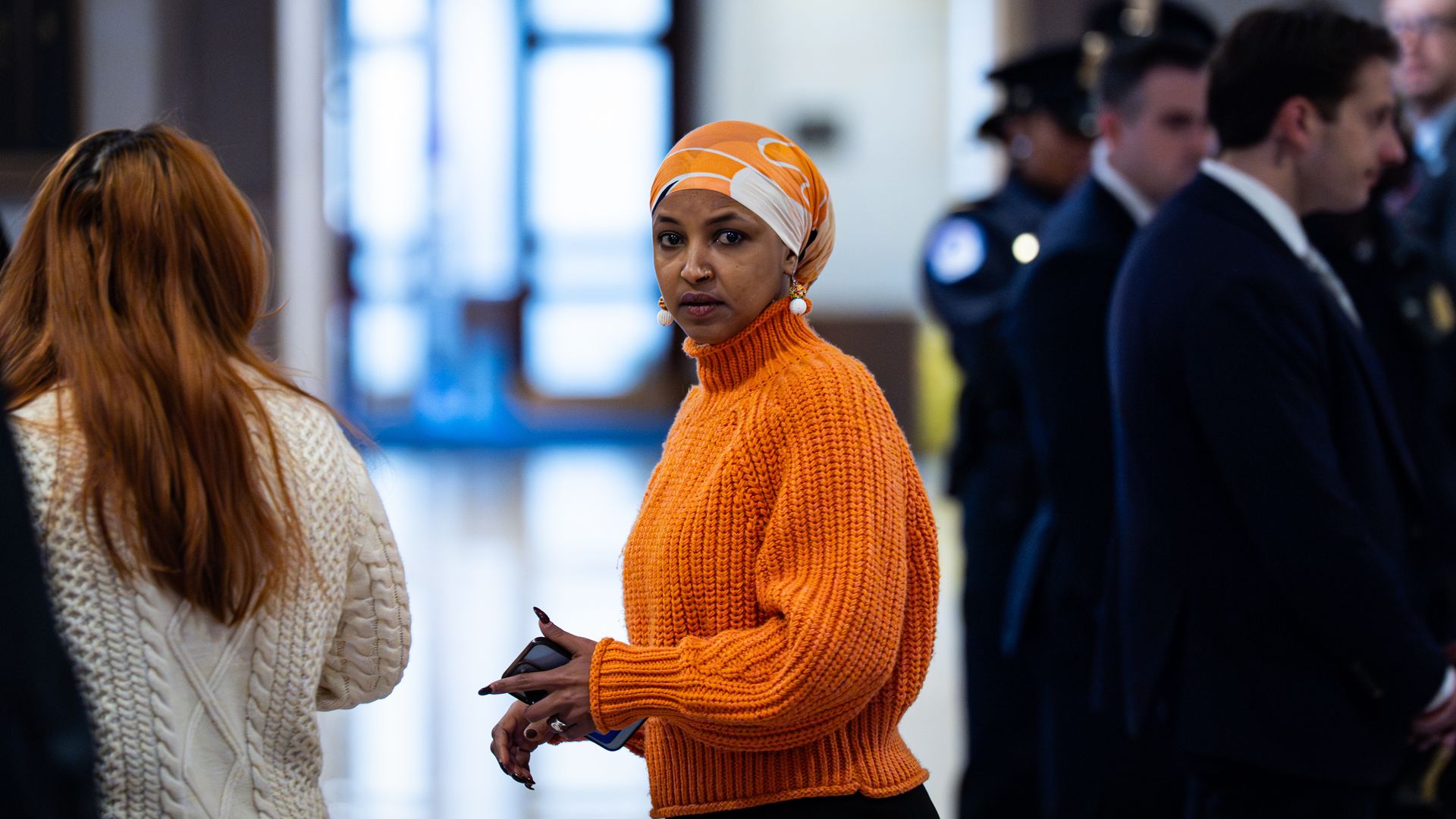 Woman in an orange knit sweater and matching headscarf stands looking at the camera, holding a phone, in a busy indoor setting with blurred people and officials in the background.