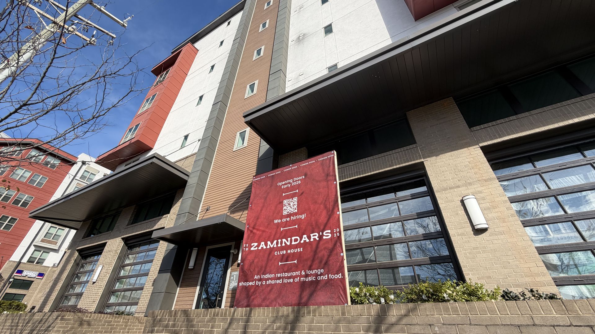 Street-level view of a multi-story modern building with tan, white, and red sections under a clear blue sky. A large red sign reads "ZAMINDAR'S CLUB HOUSE" announcing an Indian restaurant opening early 2026.
