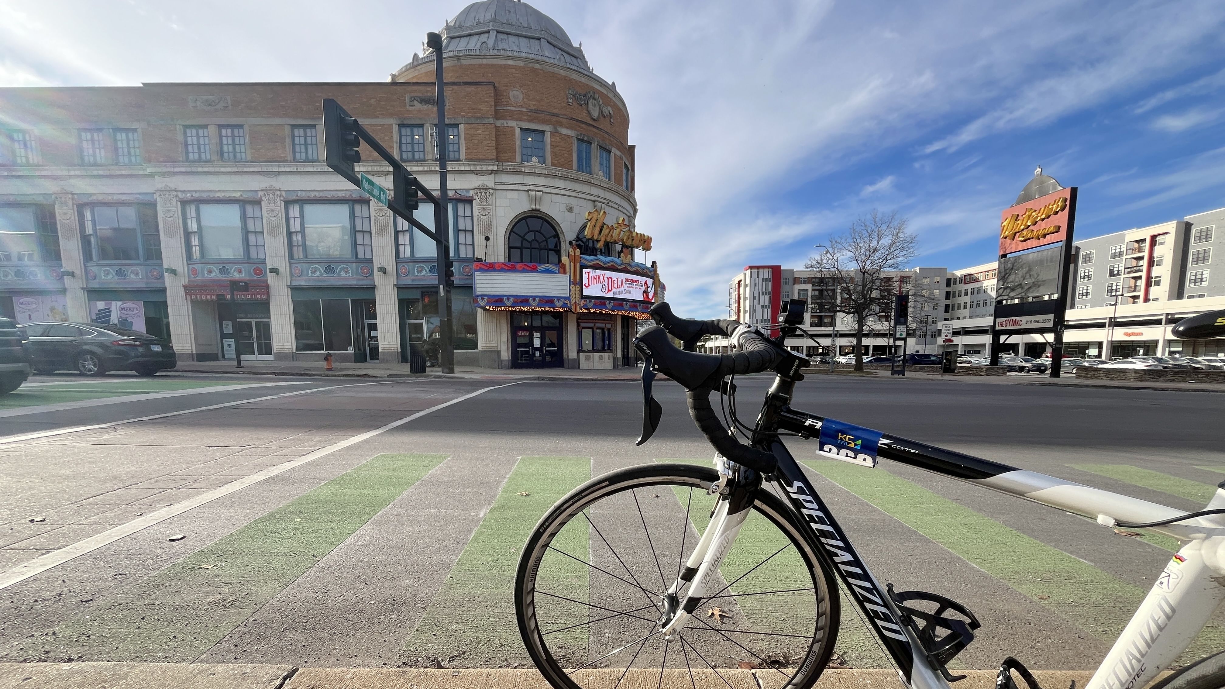 Black and white Specialized road bike parked in a green bike lane facing a large historic brick and white building with a dome and a sign saying "Uptown Theater" under a partly cloudy blue sky.