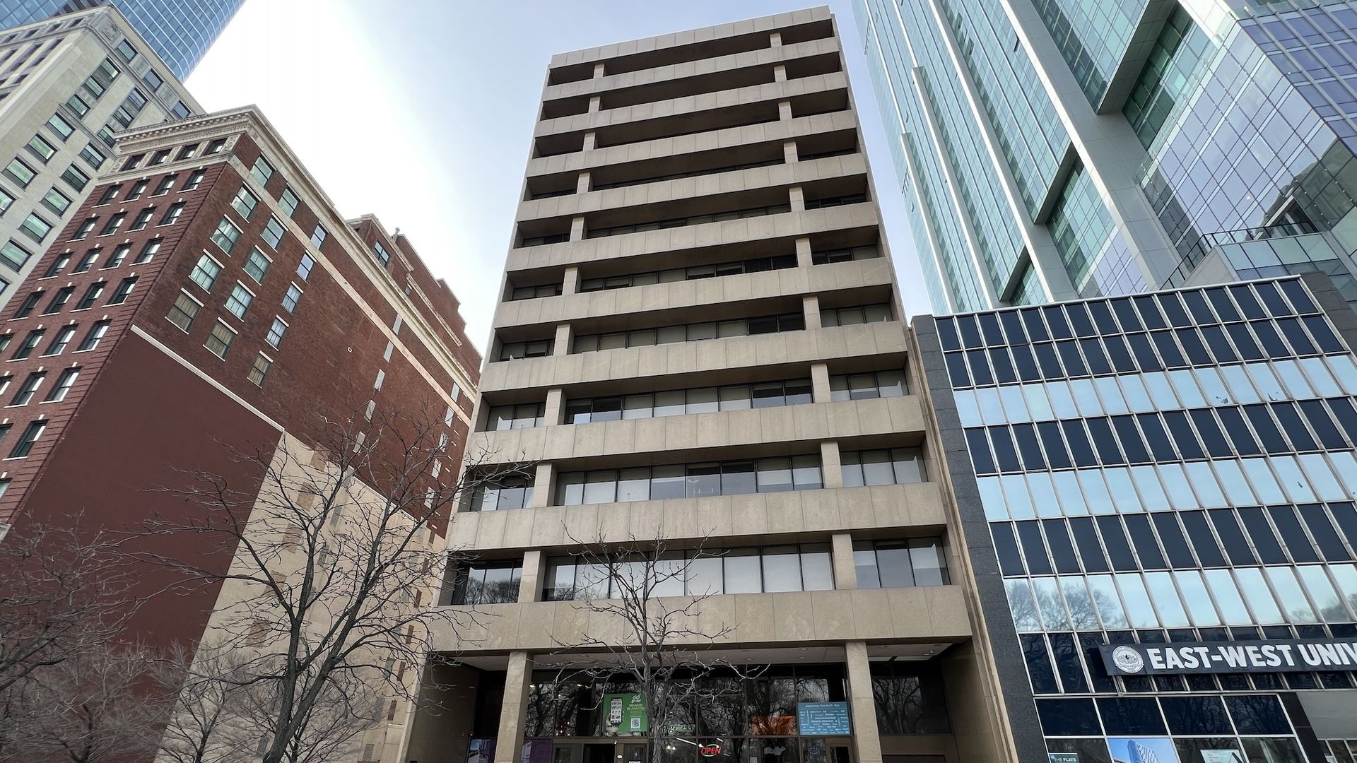 Front view of a tall beige concrete building with large windows, leafless trees, and adjacent modern glass buildings including one with the sign "EAST-WEST UNIVERSITY".