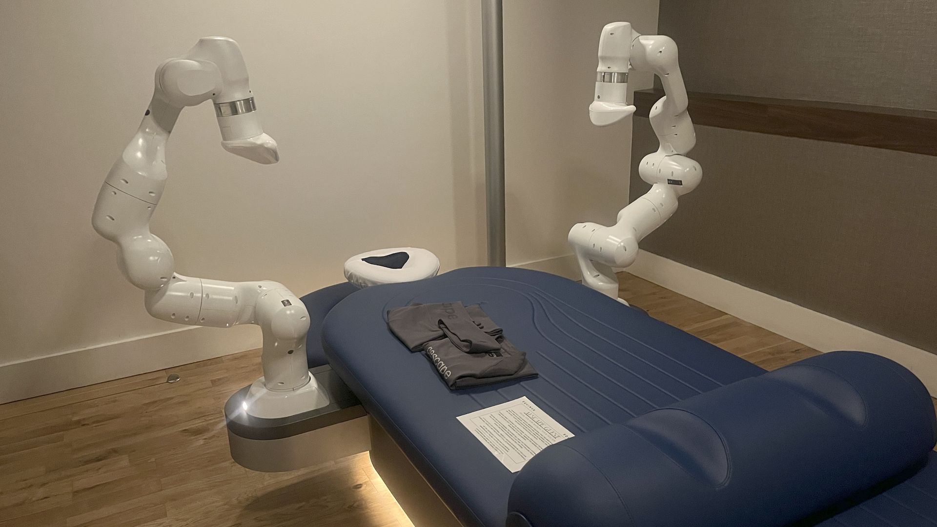 A modern blue massage table with two white robotic arms positioned on either side in a minimalistic room with wooden floor and neutral walls.