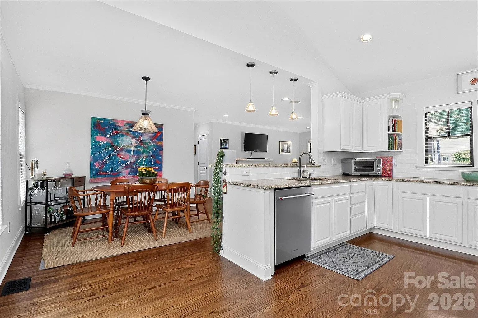 Bright open-plan kitchen and dining area with white cabinets, granite counters, and a central island. Wooden dining table, colorful abstract painting, bar cart, and pendant lights.